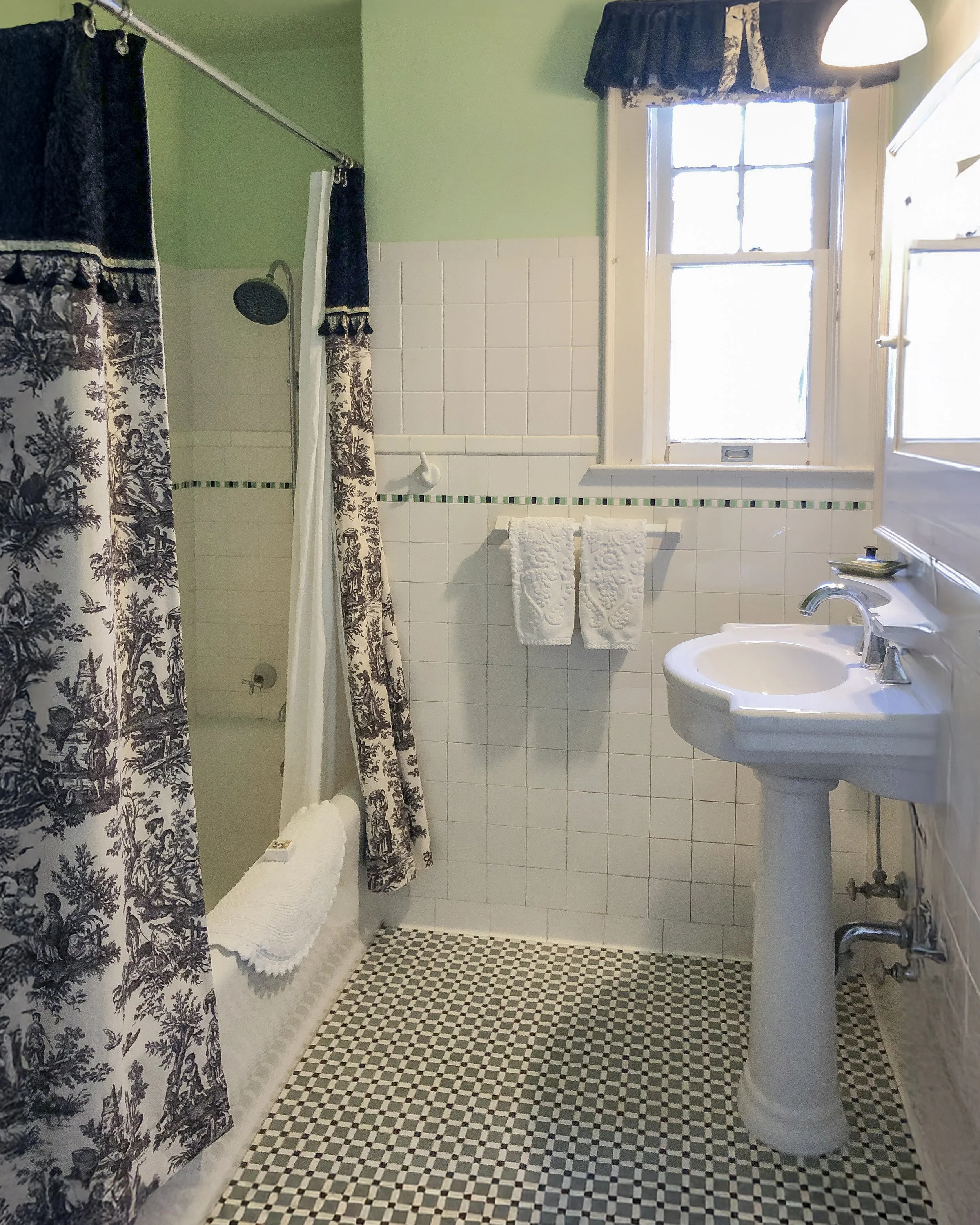 Vintage bathroom with pedestal sink, patterned shower curtains, and black-and-white checkered floor tiles. The room has white tiles on the walls, a window with curtains, and towels hanging beside the sink.