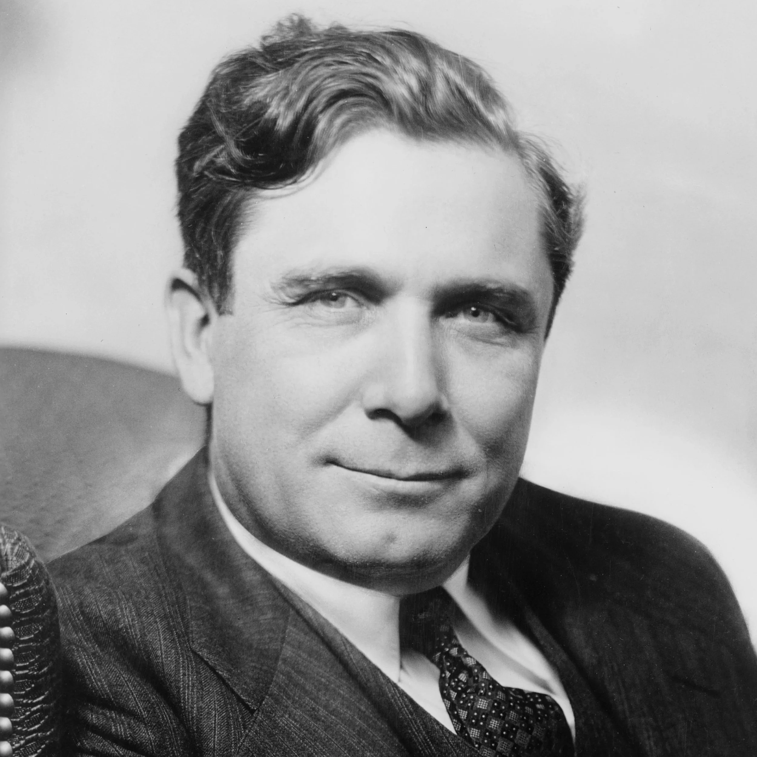 Black and white portrait of a man with neatly combed hair, dressed in a suit, sitting in a chair with a faint smile.