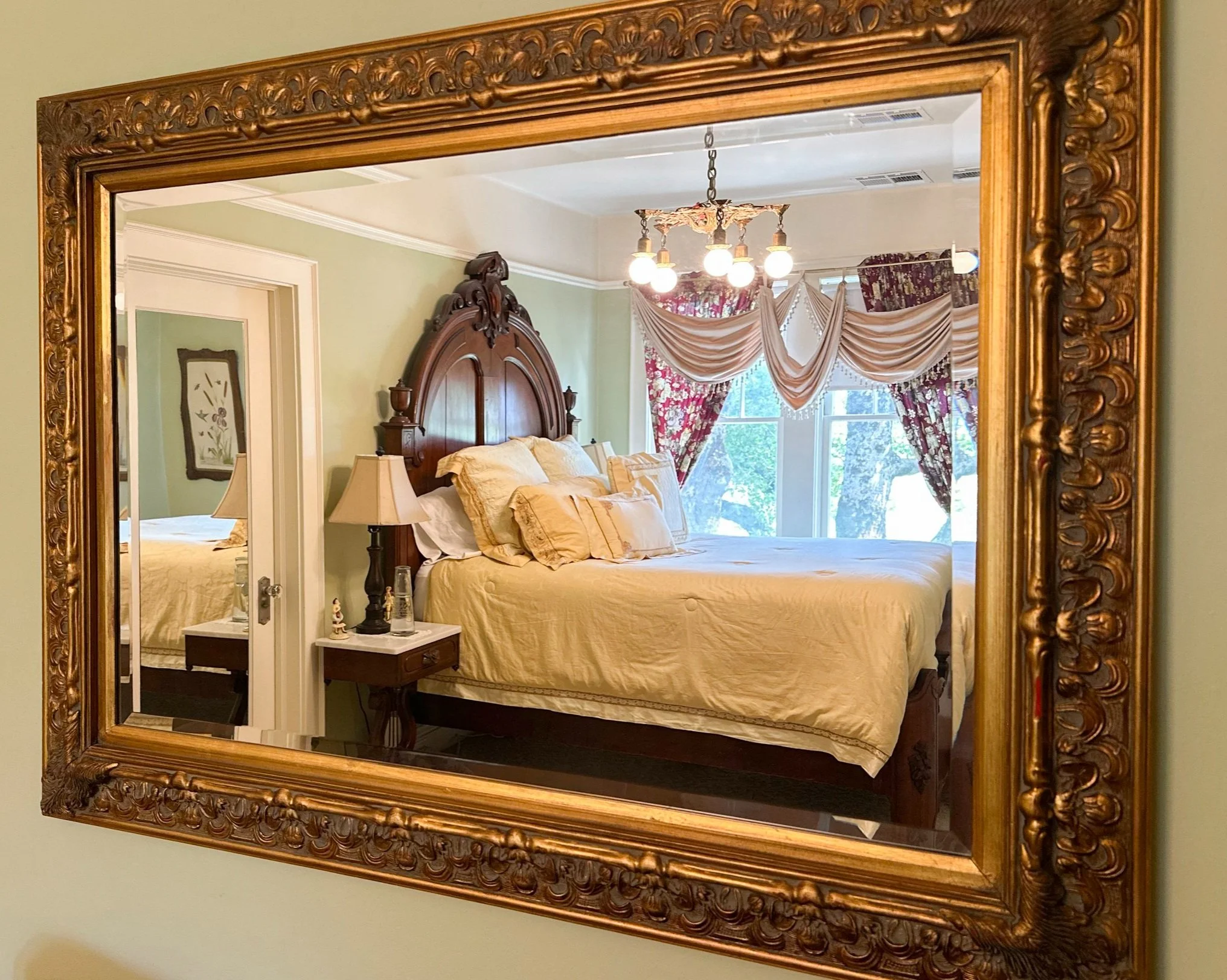 A large mirror with an ornate gold frame reflects a bedroom featuring a wooden bed with a high sculpted headboard, cream bedding, matching pillows, a nightstand with a lamp, and a window with floral curtains and draped valances. A chandelier with exposed bulbs hangs from the ceiling.