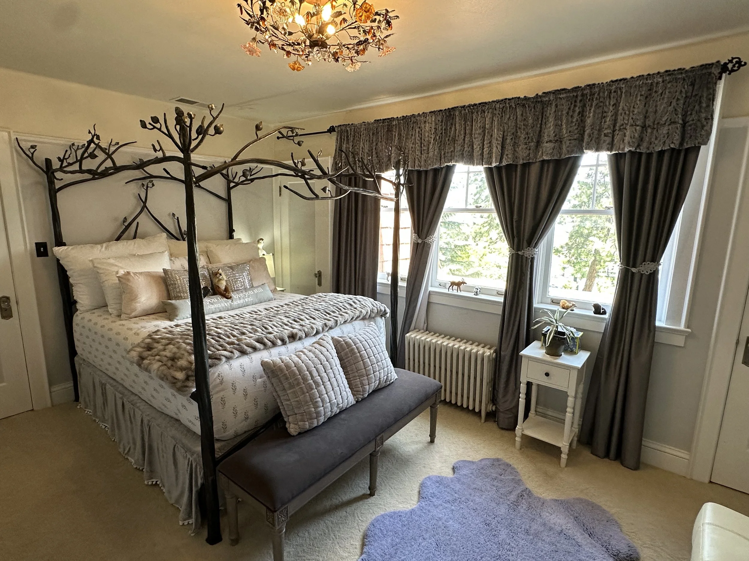 Bedroom with a tree-themed bed frame, decorative pillows, gray curtains, a chandelier, and a small side table with a plant.