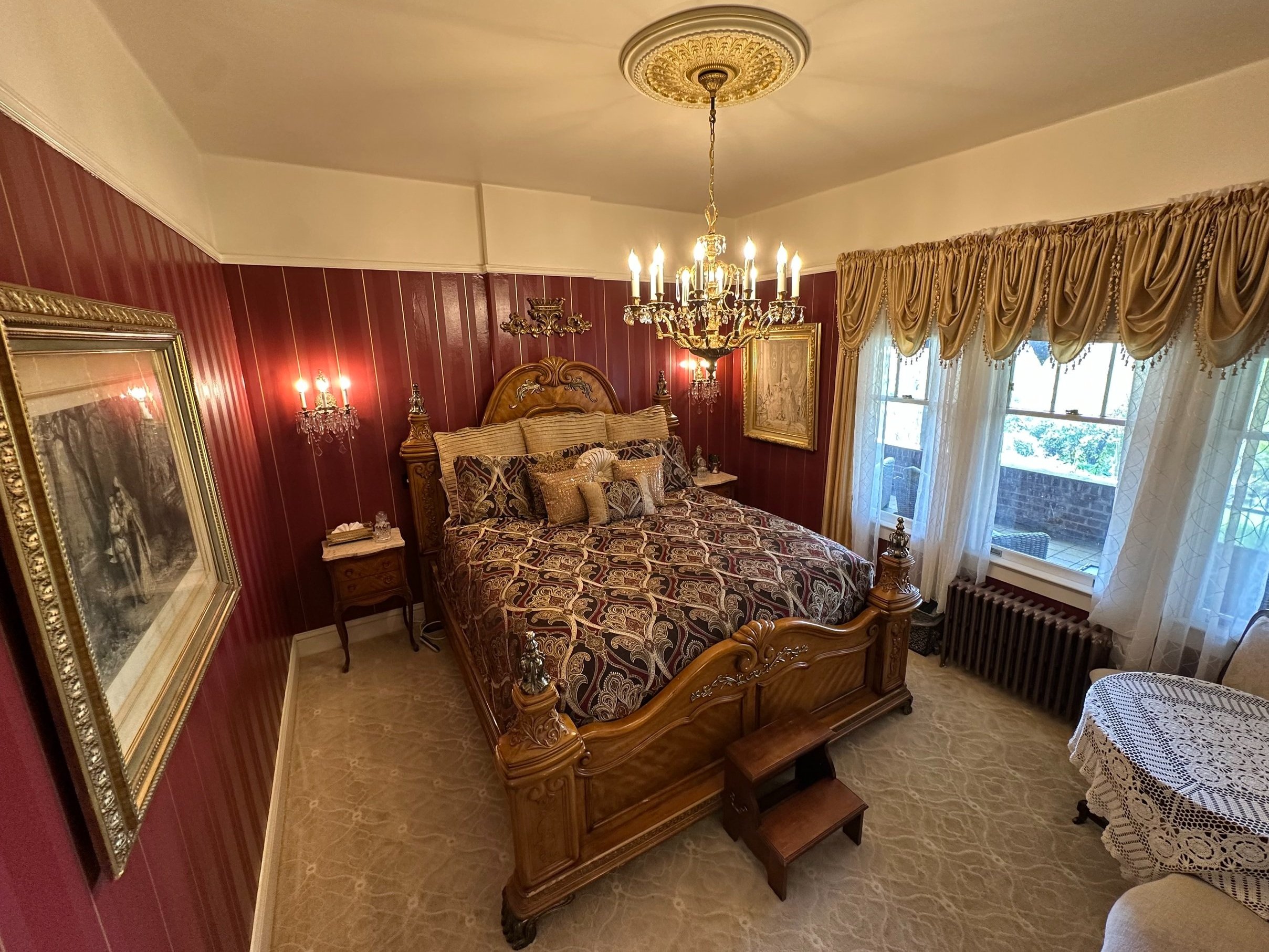 A luxurious bedroom with vintage wooden bed, ornate chandelier, red and gold decor, gold curtains, lace tablecloth, and classical artwork.