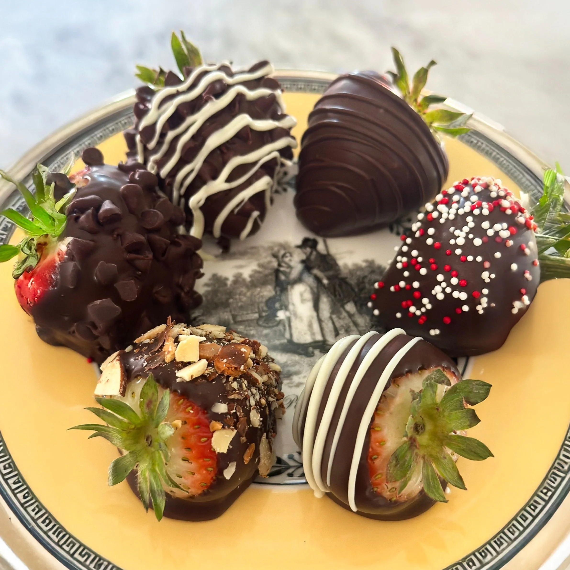 A variety of chocolate-covered strawberries on a decorative plate. Some are drizzled with white chocolate, sprinkled with nuts, or decorated with red and white sprinkles.
