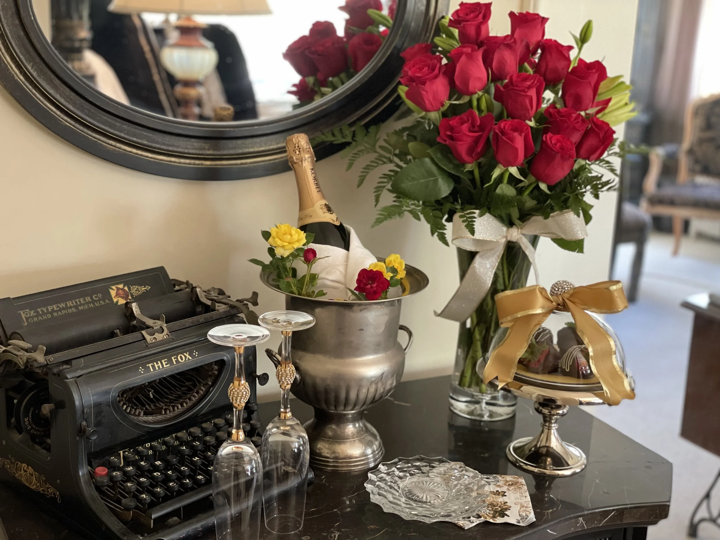 Vintage typewriter with "THE FOX" branding, two empty wine glasses, bottle of Korbel champagne in an ice bucket beside red and yellow flowers, bouquet of red roses in a vase, small cake stand with a gift bow, and decorative items on a black marble table.