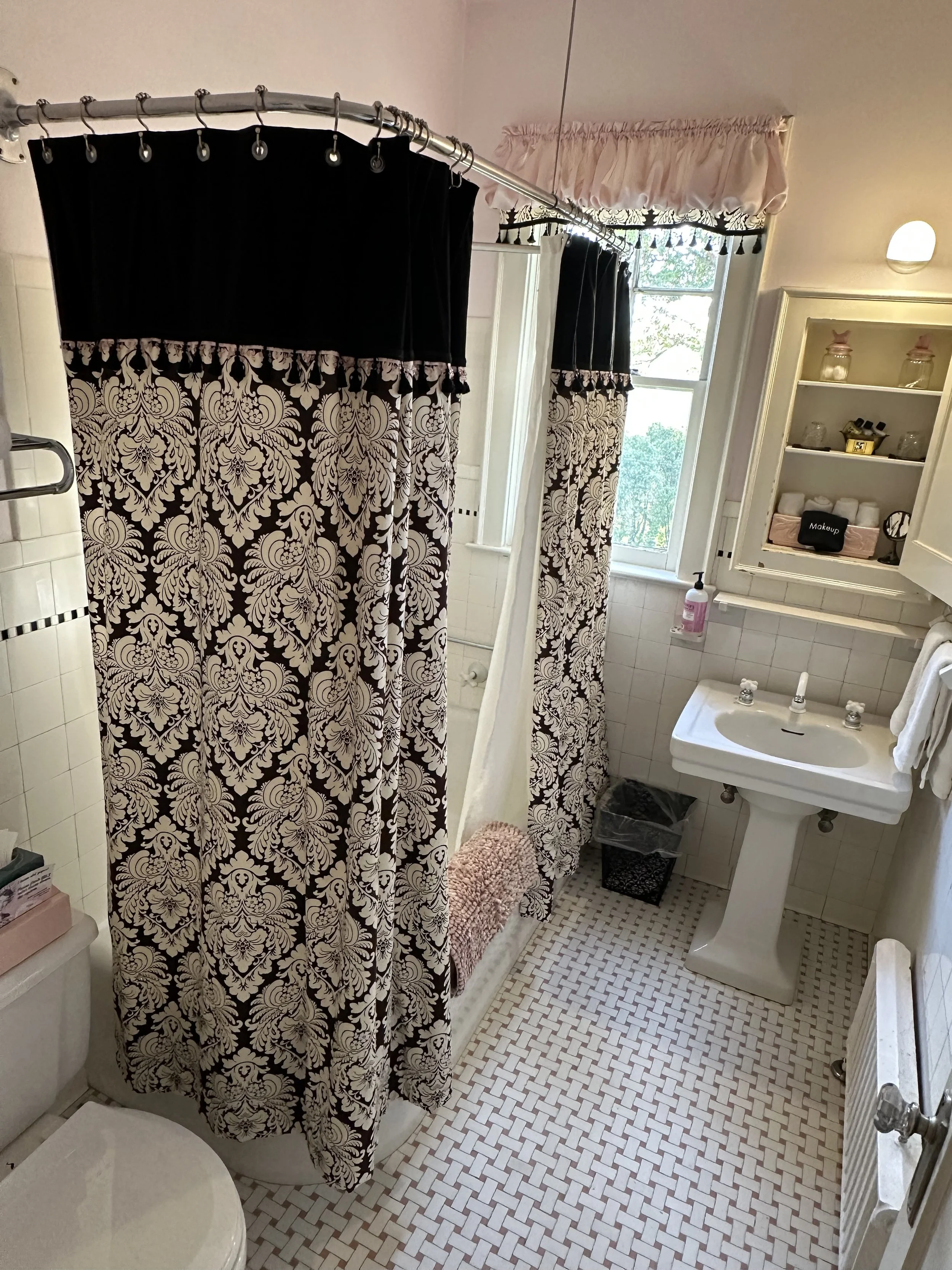 Bathroom with black and white patterned shower curtain, pedestal sink, white tile floor, shelving with toiletries, and window with valance.