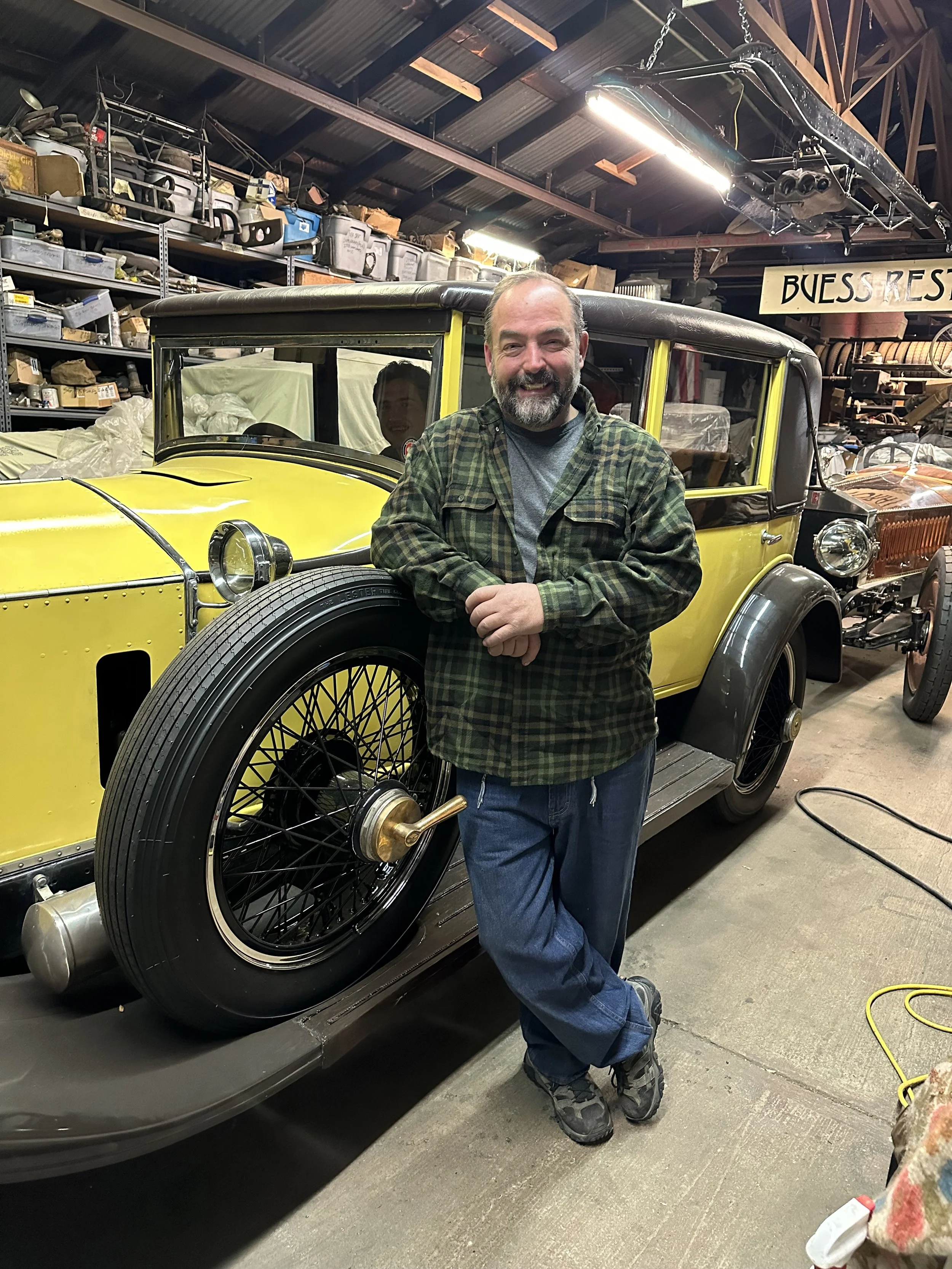 Fred Buess leaning against a vintage yellow car in a cluttered garage with shelves of tools and supplies.