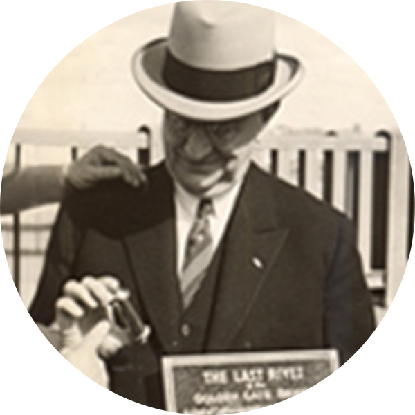 Vintage black and white photo of a man in a hat and glasses, holding a plaque labeled 'The Last Rivet,' with people gesturing nearby.