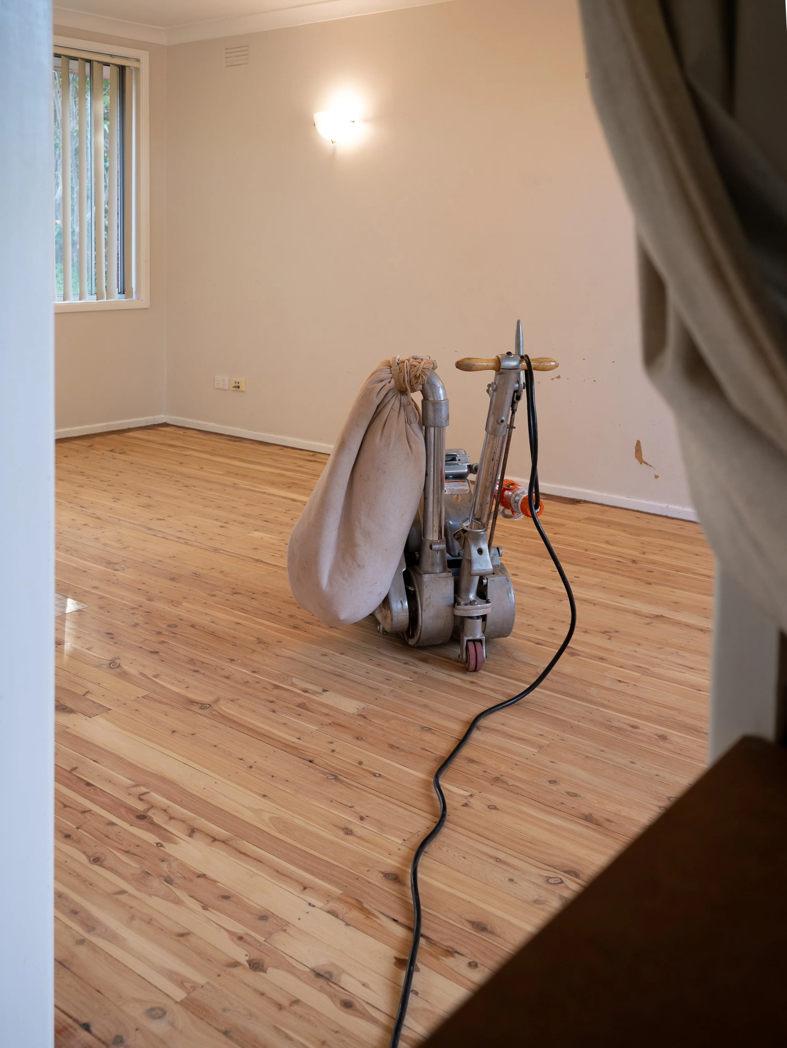 Empty room with wooden floor being varnished, white kitchen cabinets, a ceiling fan, and a floor sander in progress.