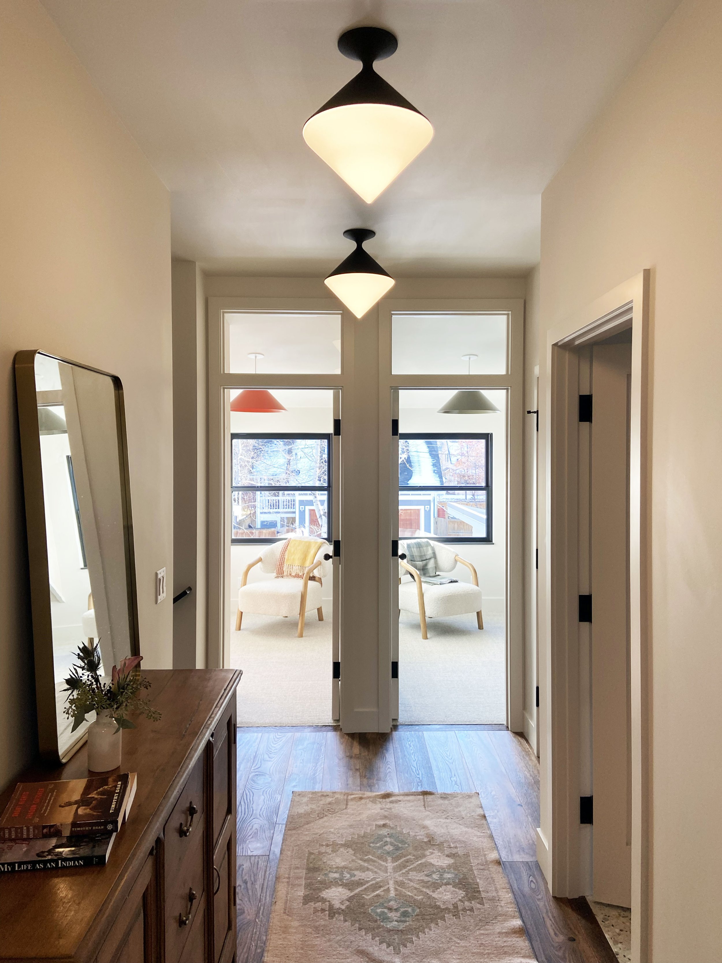 A hallway with wooden flooring and a patterned rug leading to a brightly lit room. The hallway has two modern pendant lights on the ceiling and a wooden console table on the left with books and a small vase of flowers. At the end, there is a glass door opening to a room with two armchairs and a large window, allowing natural light to enter.