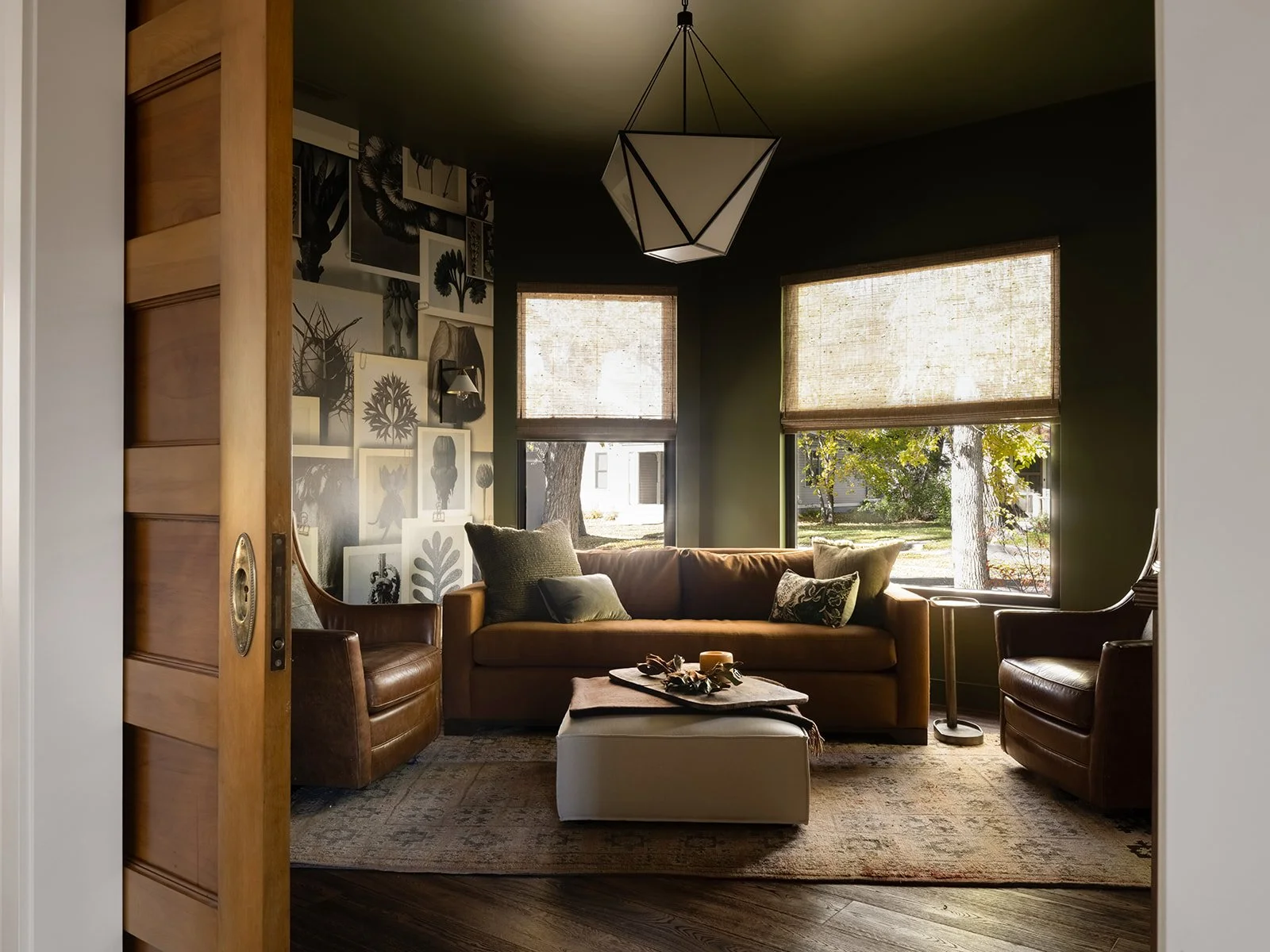 Formal library, armchair, beige ottoman on patterned rug, black and white artwork on dark green wall, large windows with beige shades, wooden sliding door, modern geometric ceiling light, and trees visible outside. Bozeman, Montana, Remodel.