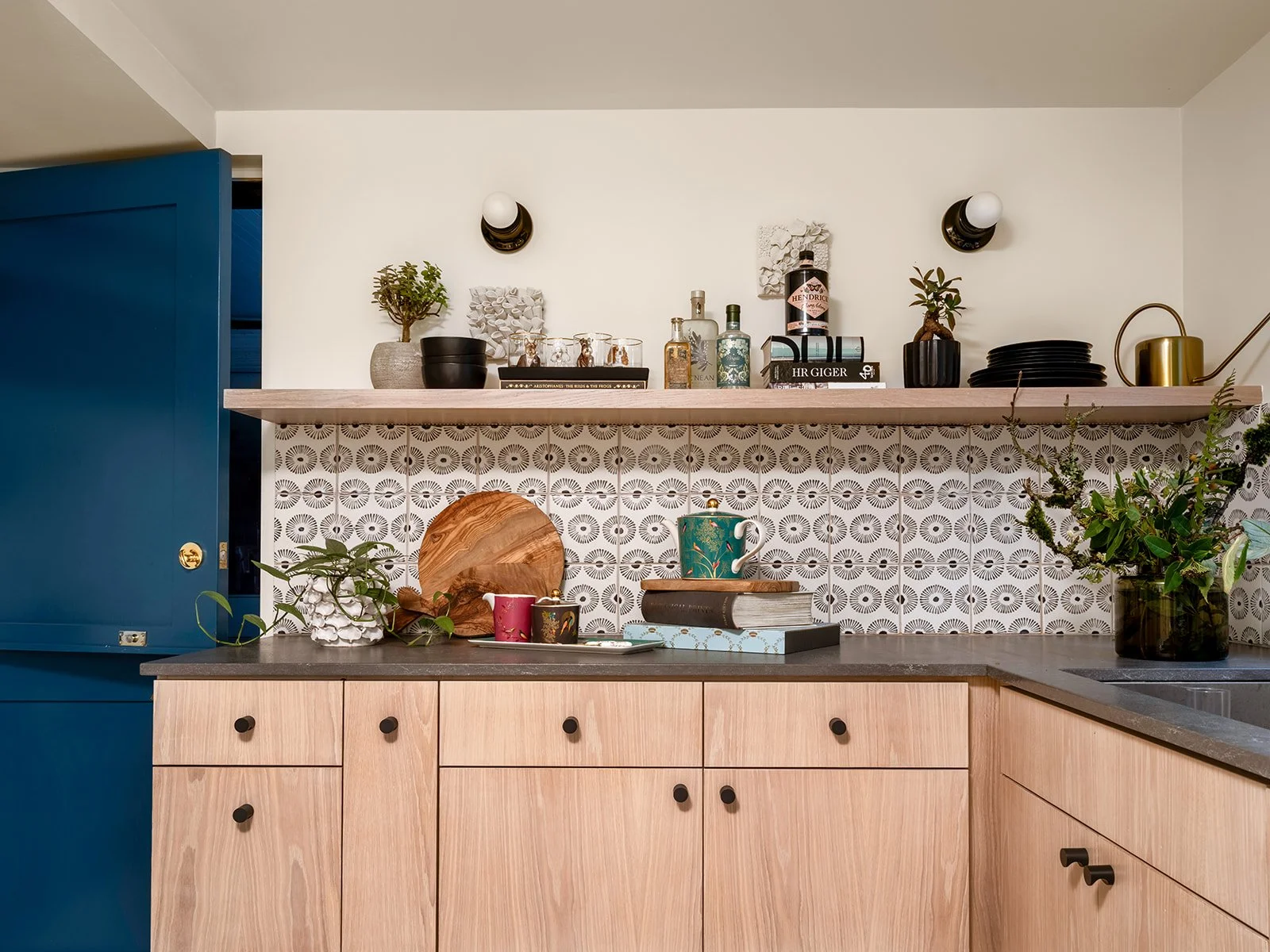 Open kitchen with wooden cabinets, black knobs, and a gray countertop. Open shelf with plants, books, bottles, and kitchenware. Back wall with patterned tiles and a large potted plant on the counter. Seattle, Queen Anne, Basement, Remodel.