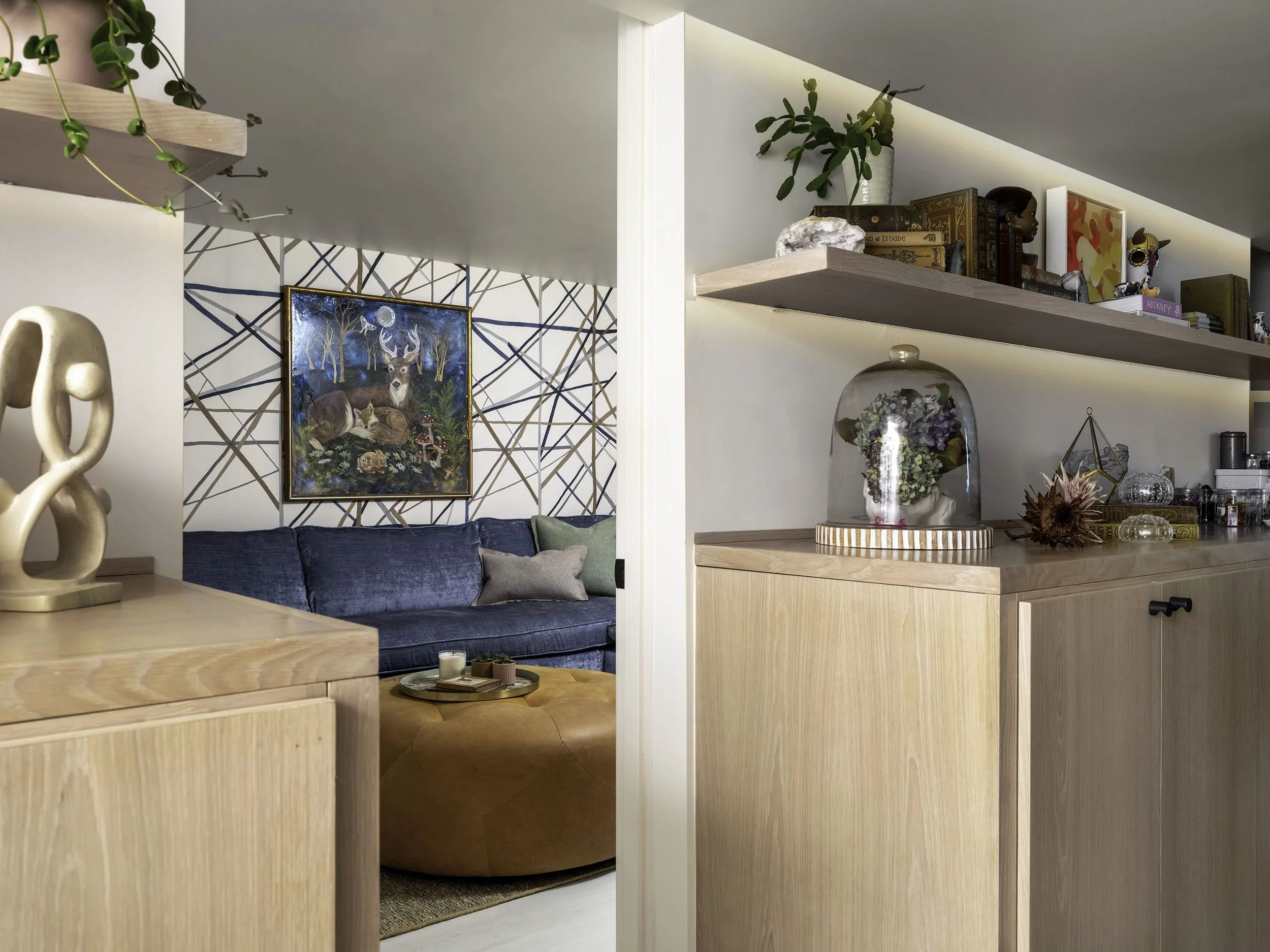 Living room with wooden cabinets, shelves with decorative items, a blue sofa, and a painting of deer on a geometric-patterned wall.