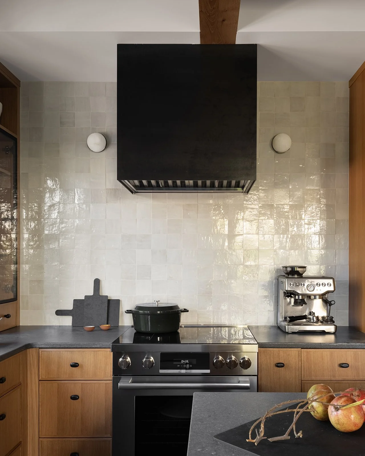 Kitchen with a stove, black pot, coffee machine, cutting boards, and apples on the counter, beige tiled backsplash, wooden cabinets, and black range hood