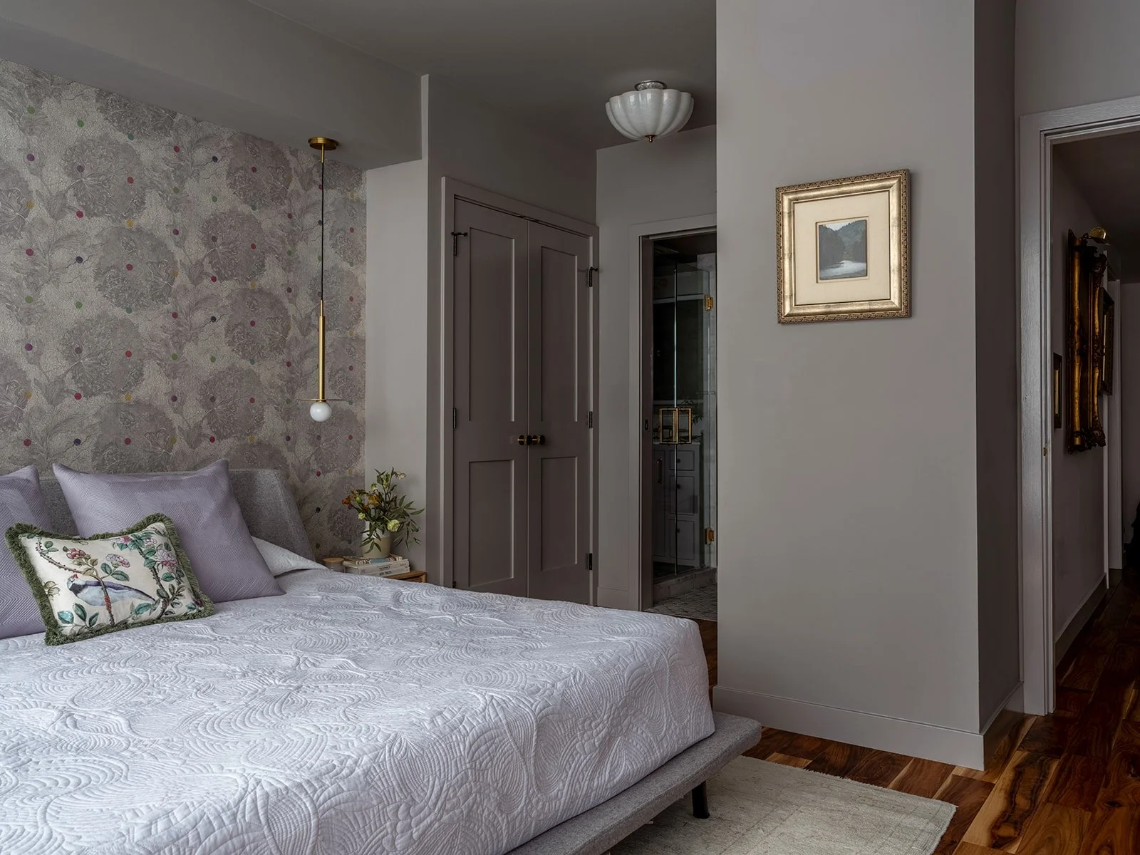 A bedroom with a gray upholstered bed, decorative pillows, a nightstand with flowers, a patterned wallpaper accent wall, a hanging pendant light, and framed art on the wall. Seattle, Belltown, Condo, Remodel.