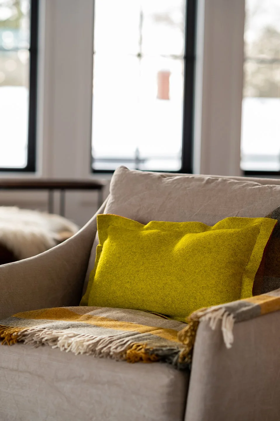 A beige sofa with a bright yellow pillow and a fringed throw blanket in a living room during daylight, with windows showing a snowy landscape outside. Seattle, Capitol Hill, Remodel.