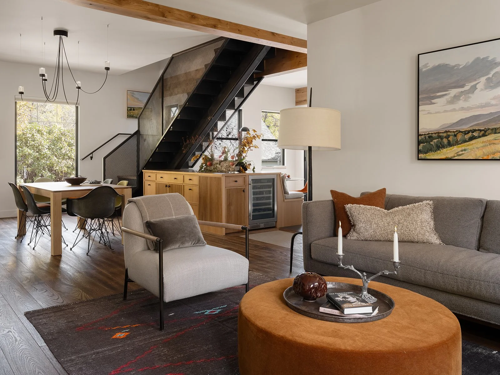 A modern living room and dining area with wooden flooring, a gray sofa with cushions, a wooden coffee table with a candle holder and books, a dining table with chairs, and a staircase with black railing and mesh panels. Bozeman, Montana, Remodel.