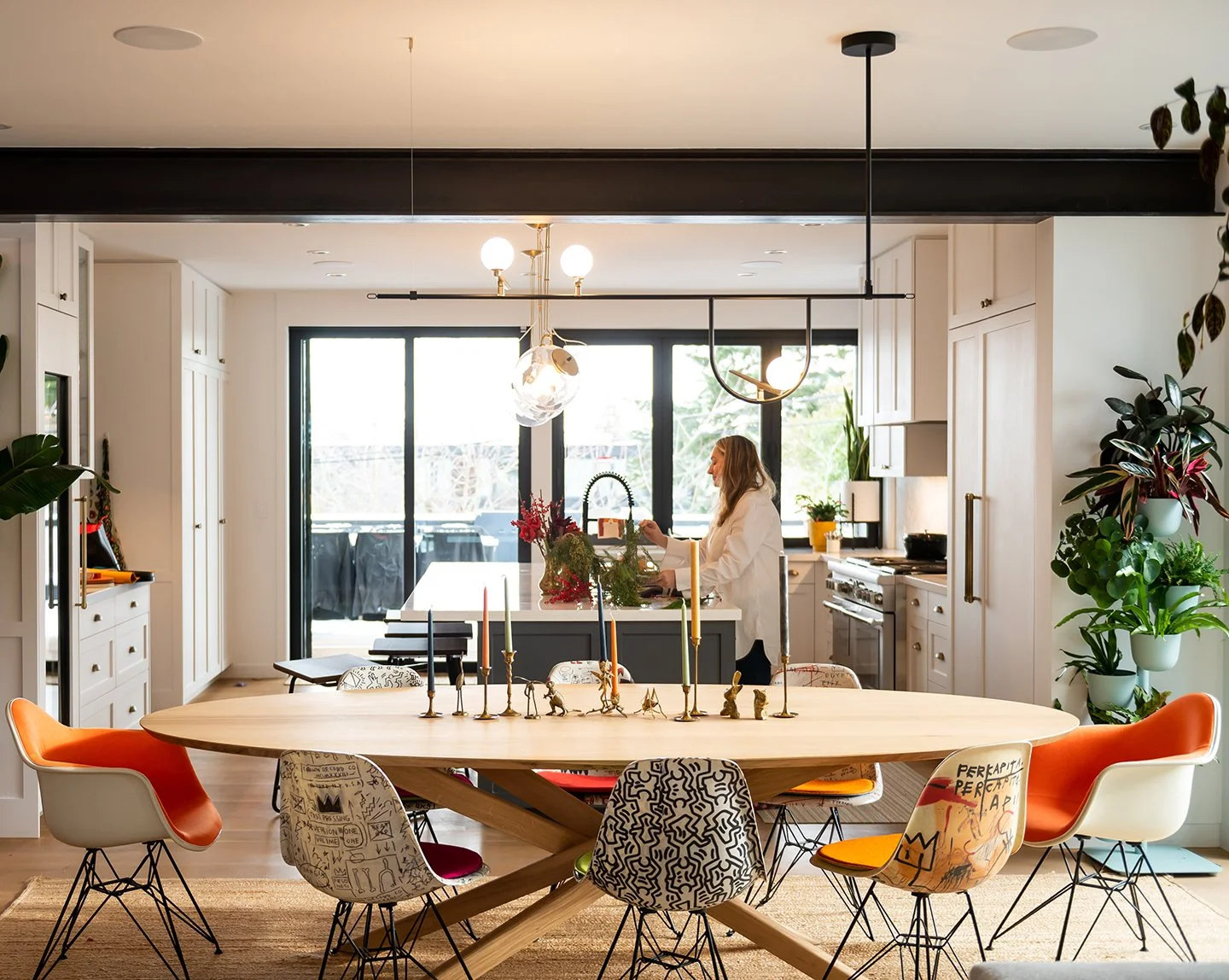 Open-concept kitchen and dining area with a large wooden table surrounded by mismatched chairs, a woman in a white blazer near the kitchen island, and large windows letting in natural light. Seattle, Capitol Hill, Remodel.