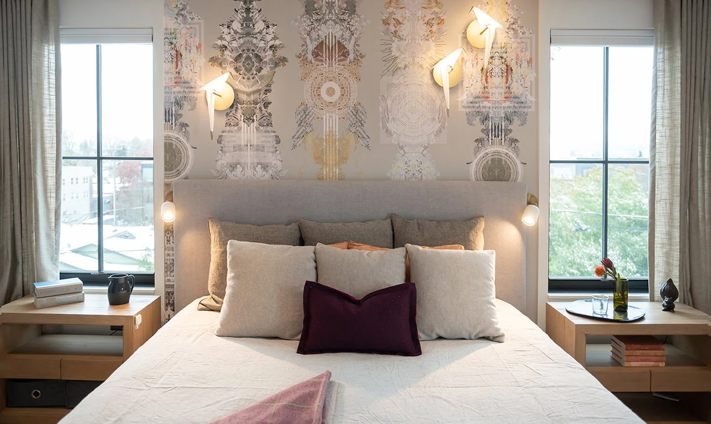 Stylish bedroom with a patterned wallpaper, two large windows, a gray upholstered bed with multiple pillows, modern wall sconces, and wooden bedside tables with books and decor.
