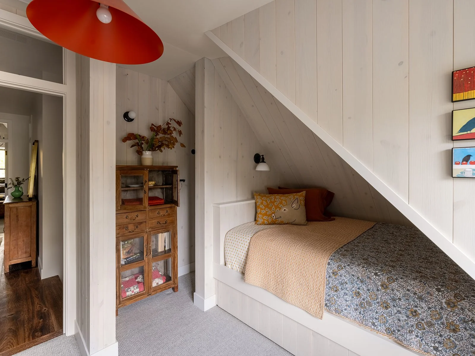A cozy bedroom with a sloped wooden ceiling featuring a bed with pillows and a bedspread, a white wall lamp, a wooden cabinet with decorations, and artwork on the wall. Bozeman, Montana, Remodel.