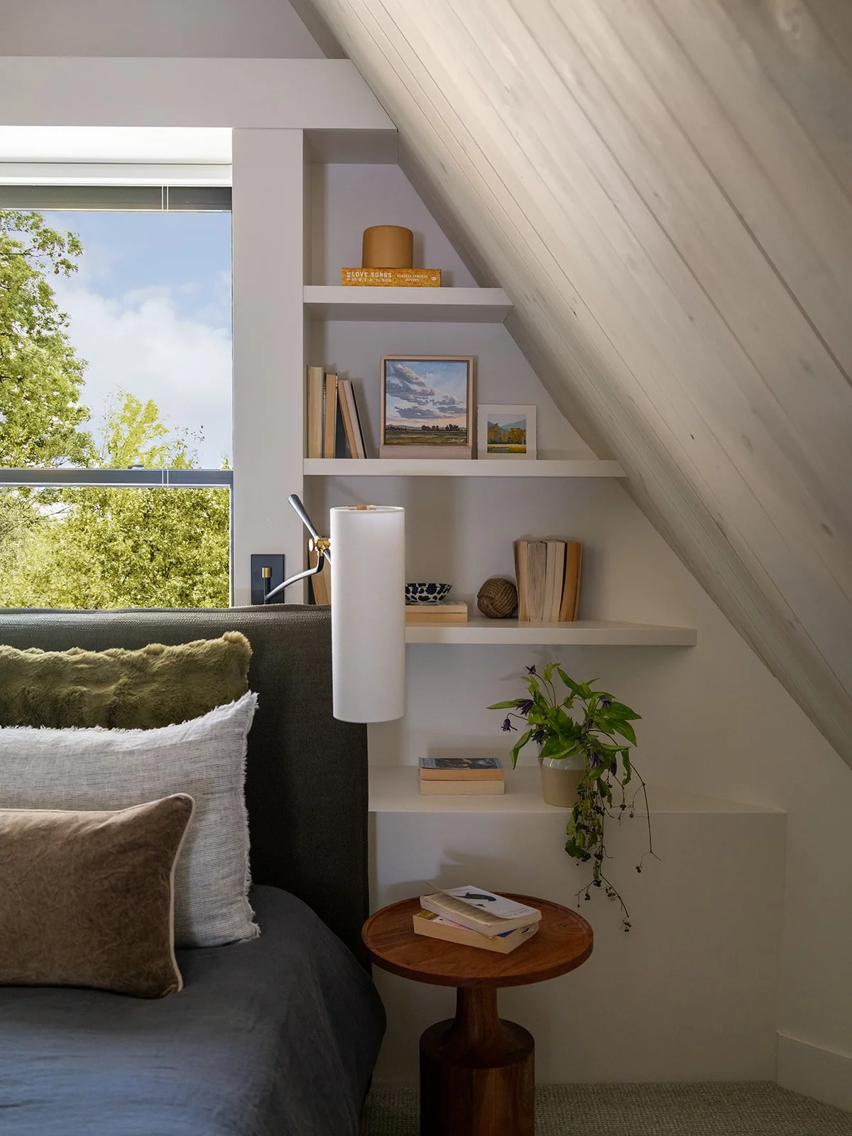 Close-up of a cozy bedroom corner with pillows, a small wooden side table with books, a potted plant, and white built-in shelves with decorative items and books beneath a sloped ceiling and a window showing trees outside. Bozeman, Montana, Remodel.
