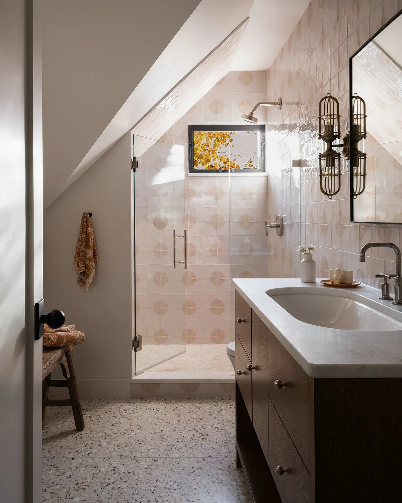 A modern bathroom with a sloped ceiling, a small window with autumn leaves outside, a glass-enclosed walk-in shower, a wooden vanity with a white countertop, and decorative wall sconces. Bozeman, Montana, Remodel.