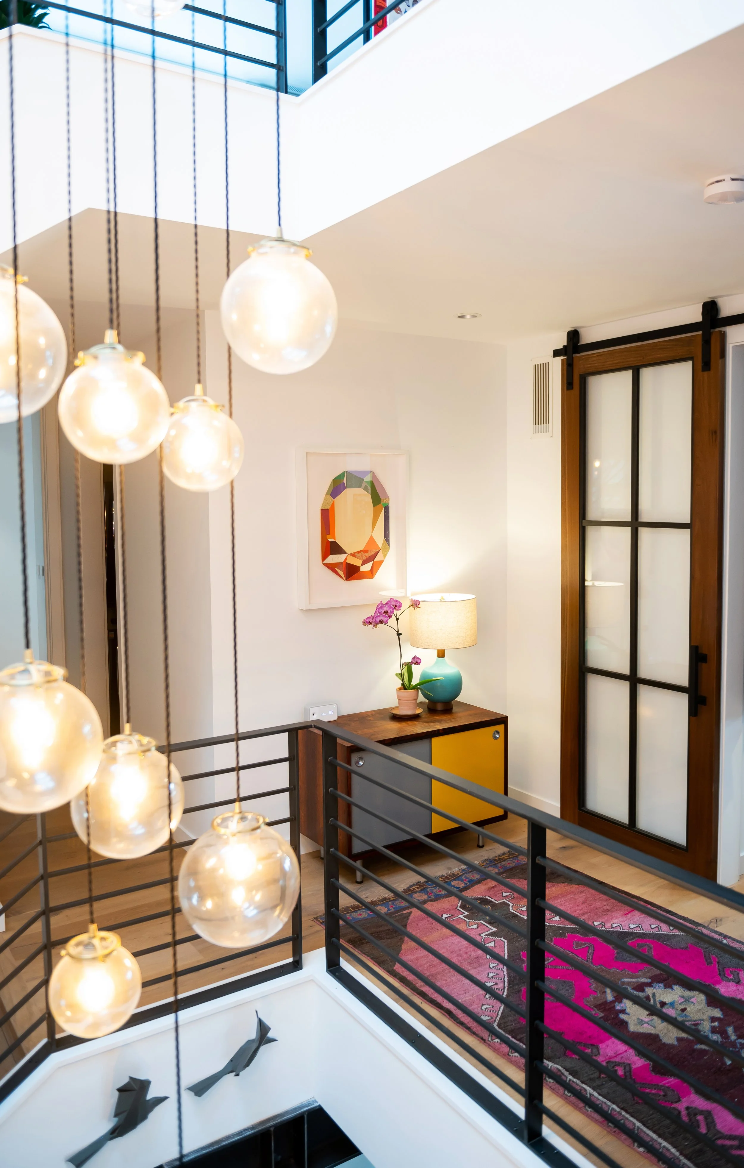 Interior view of a modern home with a staircase, hanging globe lights, a colorful rug, a yellow and wood sideboard with a lamp and potted orchid, and a sliding barn door with wooden frame and frosted glass panels. Seattle, Capitol Hill, Remodel.