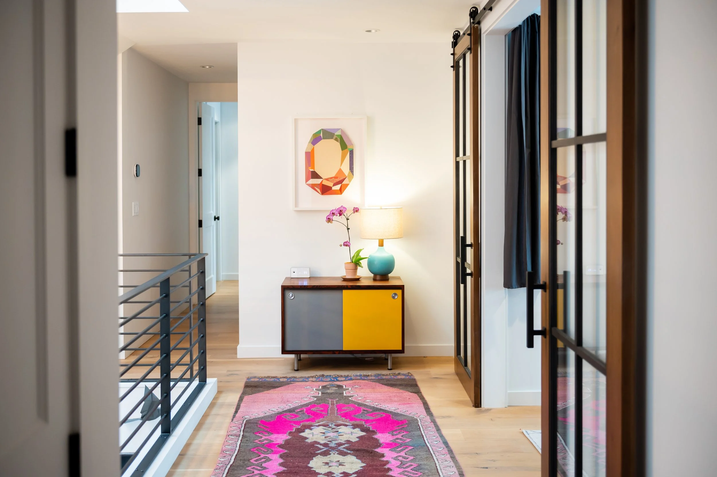 Interior view of a modern living space with a colorful patterned rug on wooden floors, a small cabinet with gray and yellow doors, a teal lamp, pink orchid, framed artwork on the wall, and glass doors on the right. Seattle, Capitol Hill, Remodel.