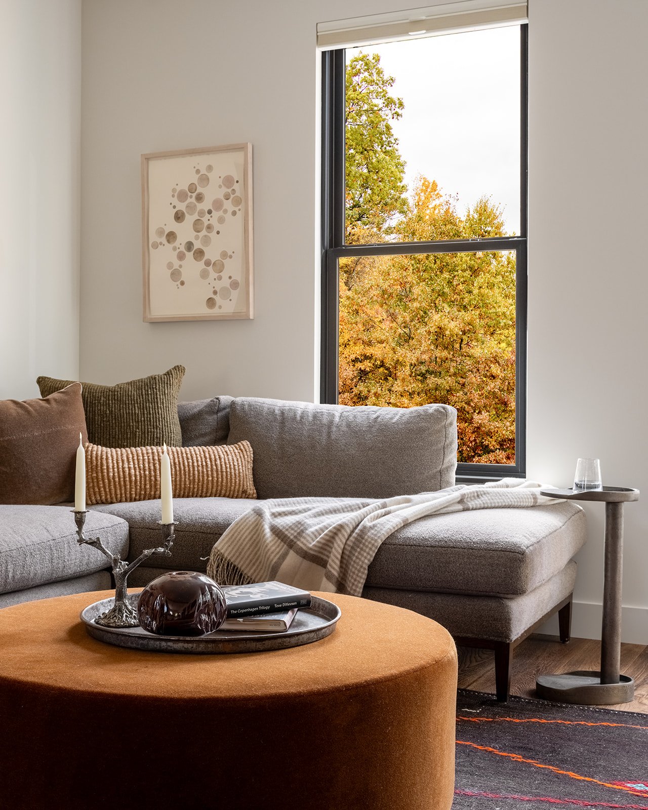 Living room with a beige sectional sofa, pillows, a wooden coffee table with a tray holding a dark decorative object and a book, a side table with a glass, a window showing autumn trees outside, and wall art. Bozeman, Montana, Remodel.