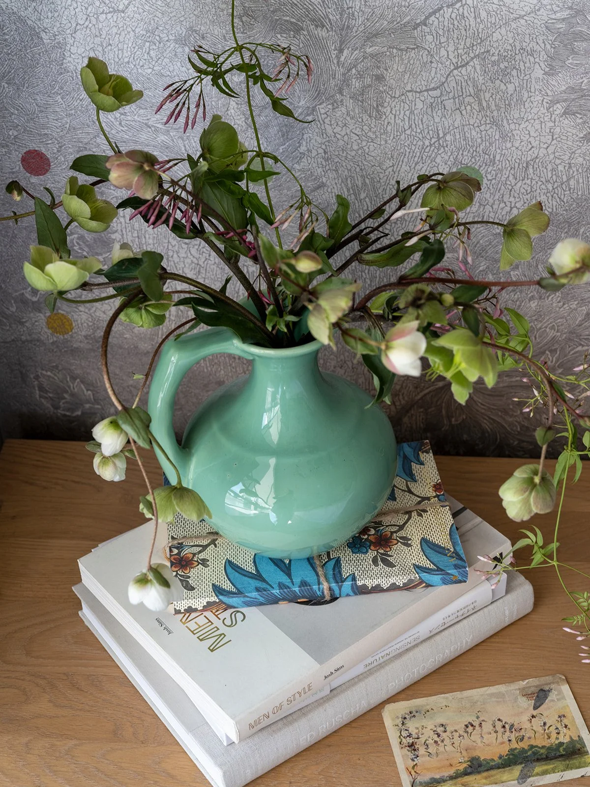A mint green ceramic vase filled with green and white flowering branches sits on top of a stack of books on a wooden surface, with a colorful cloth underneath. A decorative card is also on the surface. Seattle, Belltown, Condo, Remodel.