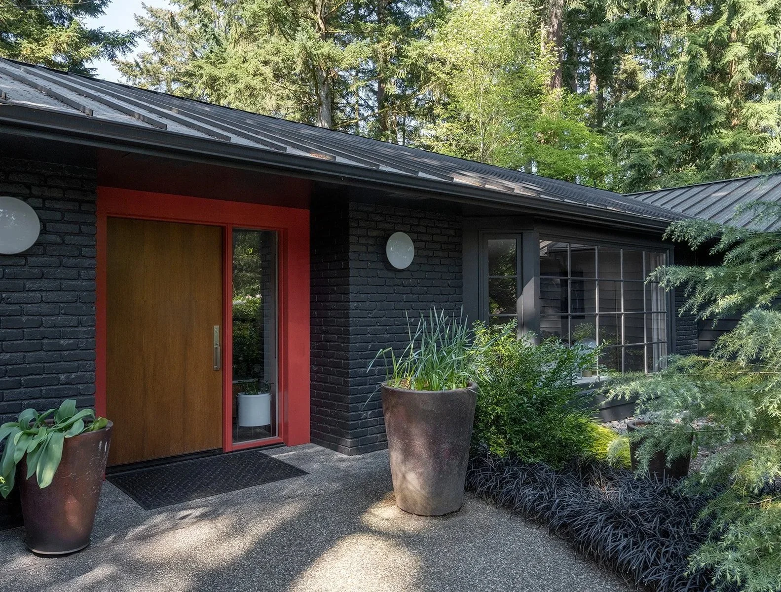 Modern house entrance with black brick walls, red trim around the door, and large potted plants, surrounded by greenery. Seattle, Mecer Island, Remodel.