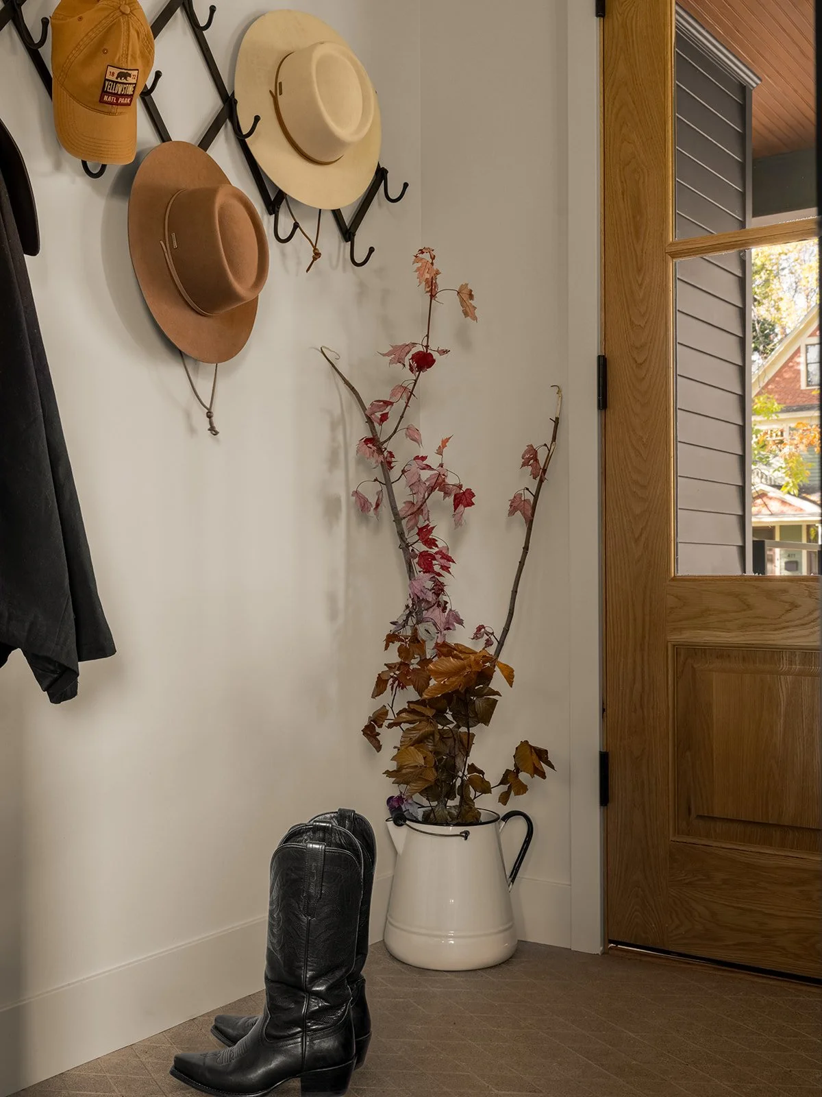 Entryway with coats and hats hanging on hooks, a large cream-colored pitcher with dried branches and leaves, a pair of black cowboy boots, and a wooden door with glass panels. Bozeman, Montana, Remodel.