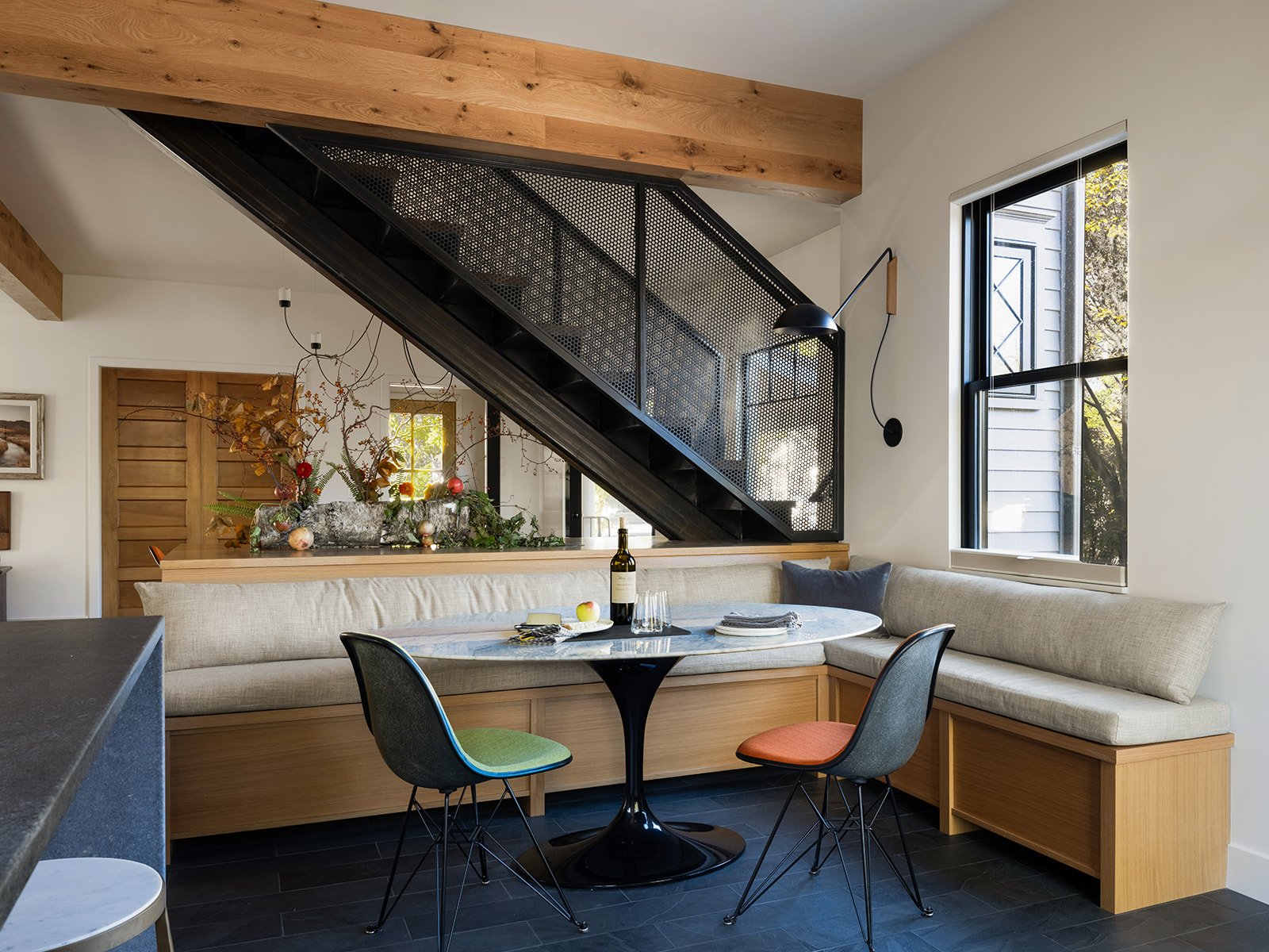 Contemporary dining area with built-in beige banquette seating, a round table, two colorful chairs, a window, and a staircase with a black metal railing and wooden accents in a modern home. Bozeman, Montana, Remodel.