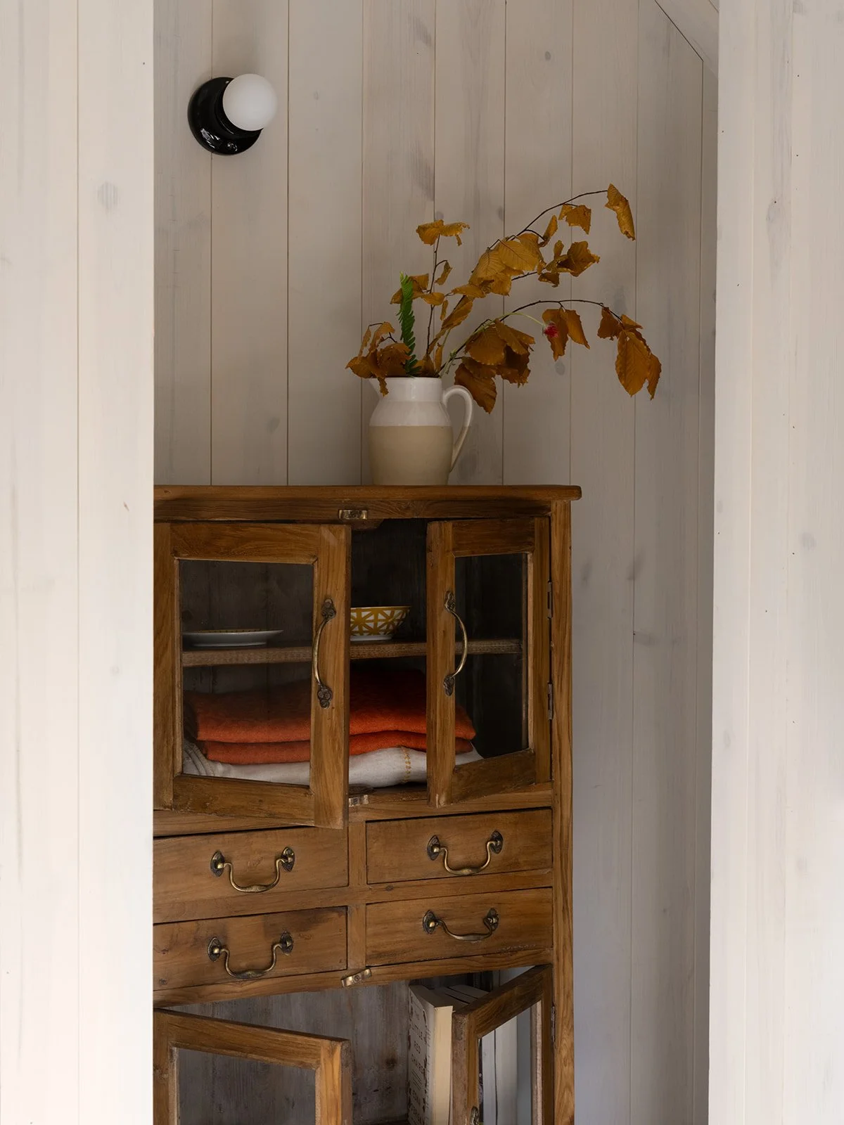 A white pitcher with dried brown leaves and a green branch on top of a wooden cabinet with glass doors and drawers, against a white wood-paneled wall. A black and white wall sconce light is mounted on the wall. Bozeman, Montana, Remodel.
