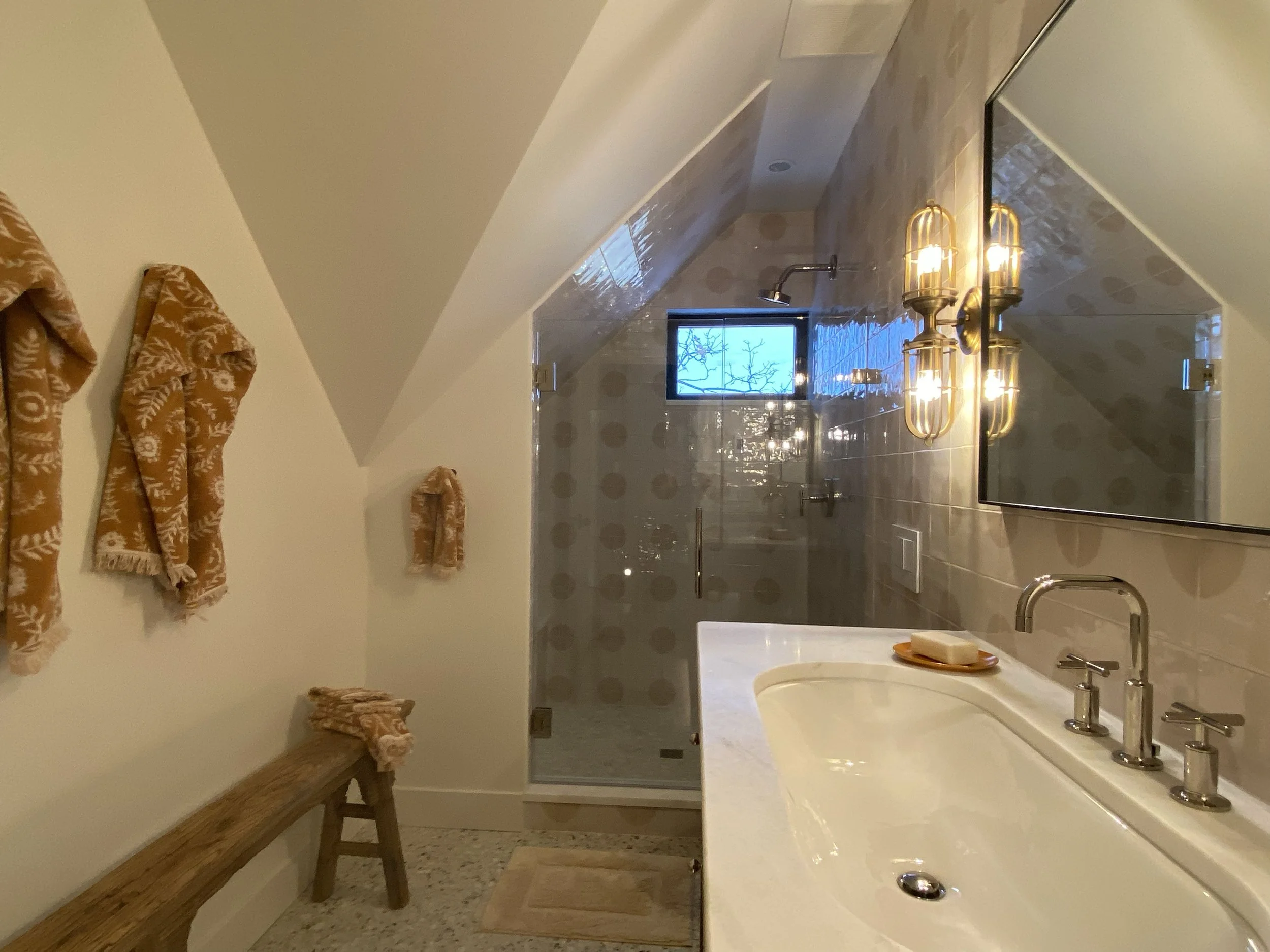 Modern bathroom with a sloped ceiling, featuring a glass-enclosed shower, white countertop with silver faucet, patterned towels, wooden bench, and wall-mounted lighting.