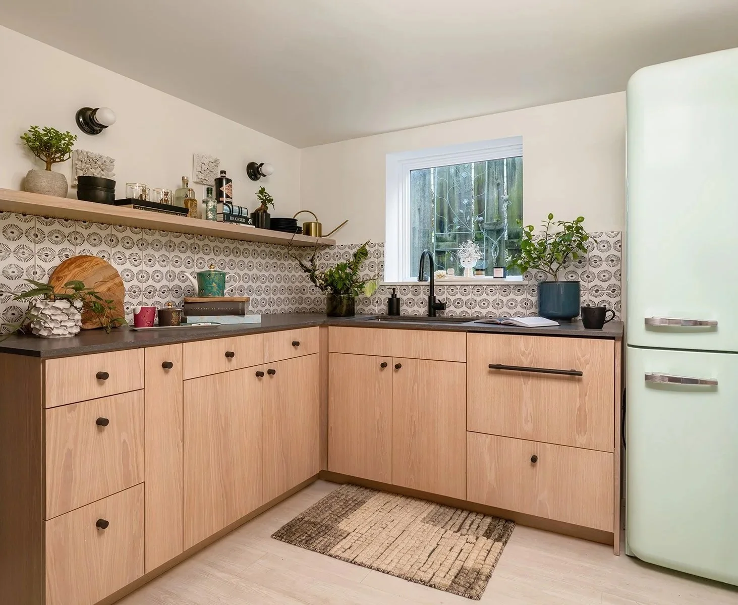 A cozy kitchen with wooden cabinets, a patterned backsplash, and a green vintage-style refrigerator. Decor items and plants are on the countertop and shelf near a window. Seattle, Queen Anne, Basement, Remodel.