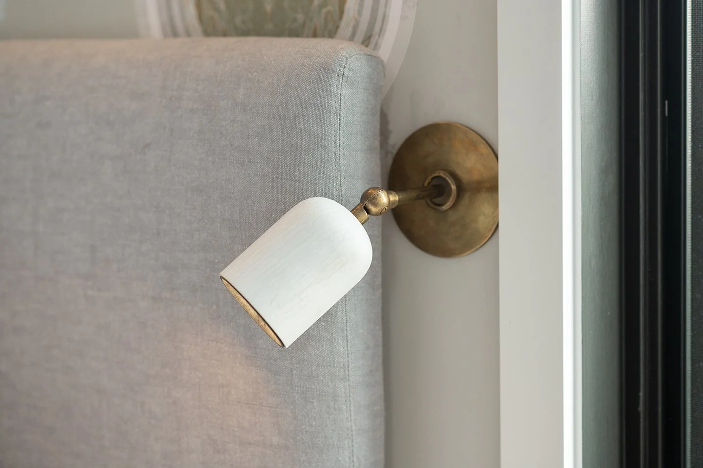 Wall-mounted reading lamp with a white lampshade directed downward, attached to a brass base on a gray upholstered headboard. Seattle, Capitol Hill, Remodel.