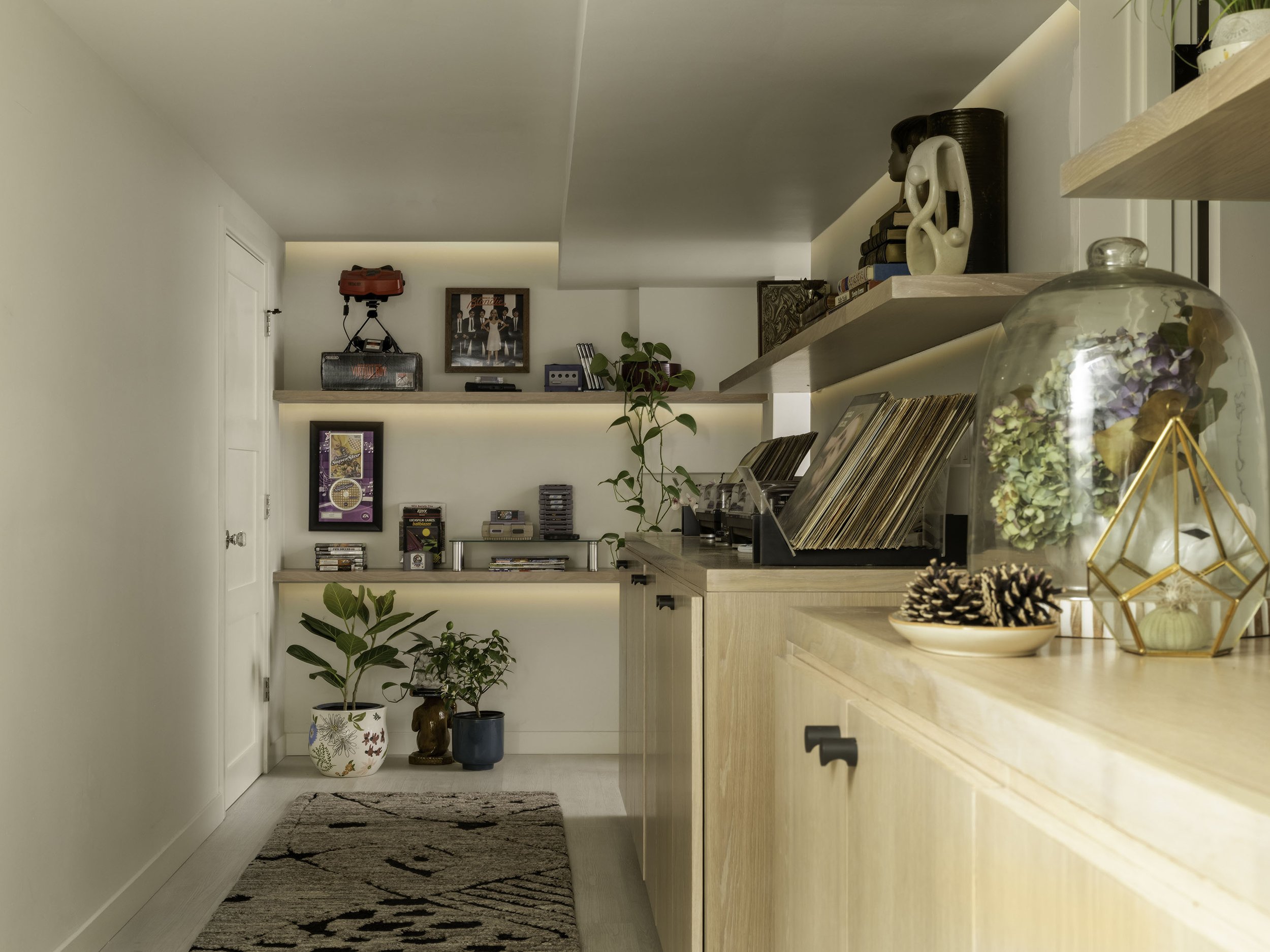 Modern interior with shelves, vinyl records, retro gaming consoles, potted plants, and decorative items on light wood furniture and cabinets.