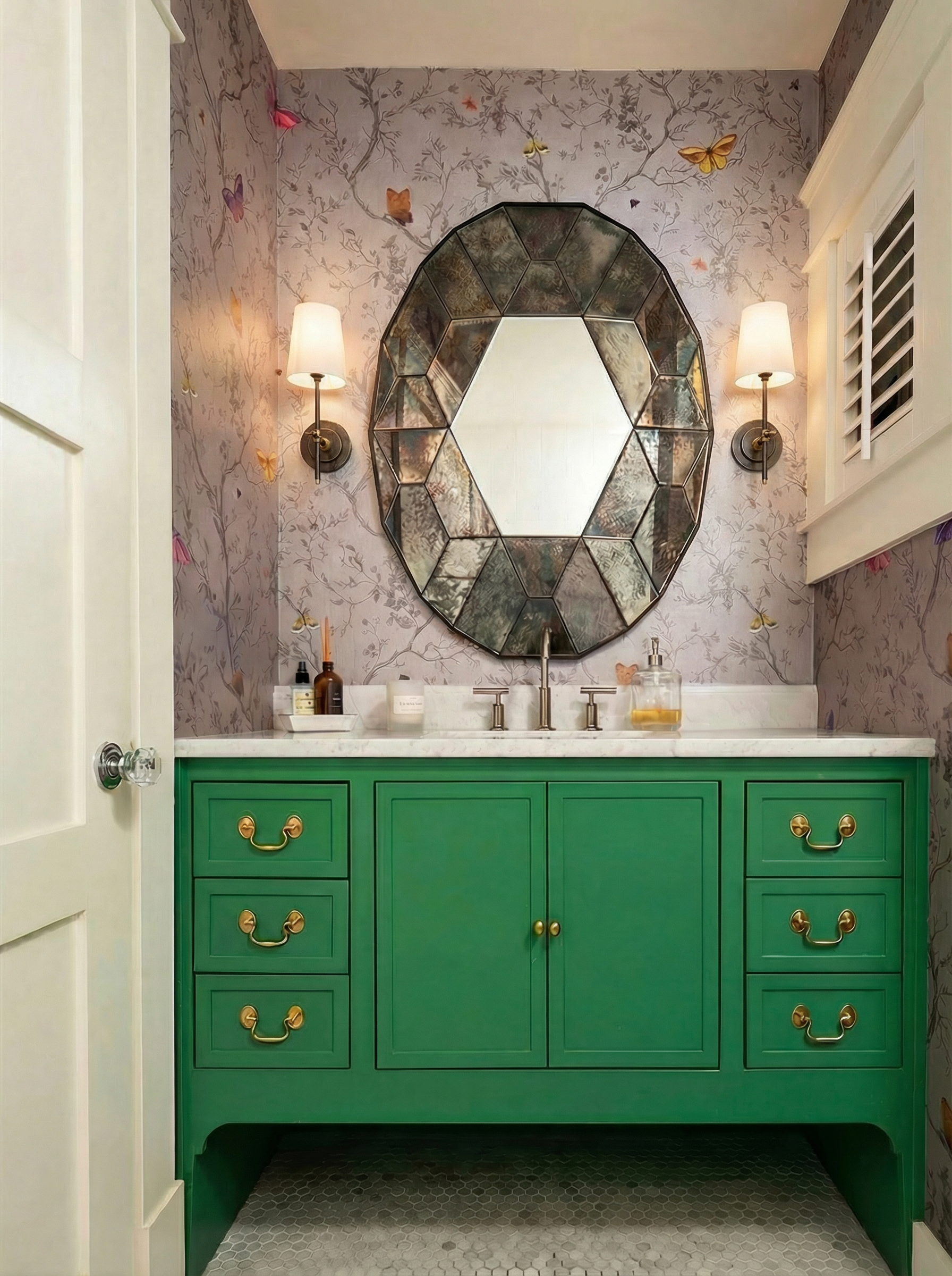 A bathroom vanity with a bright green cabinet, gold handles, and a white marble countertop, a round decorative mirror above, butterfly wallpaper, and two wall sconces with white shades.