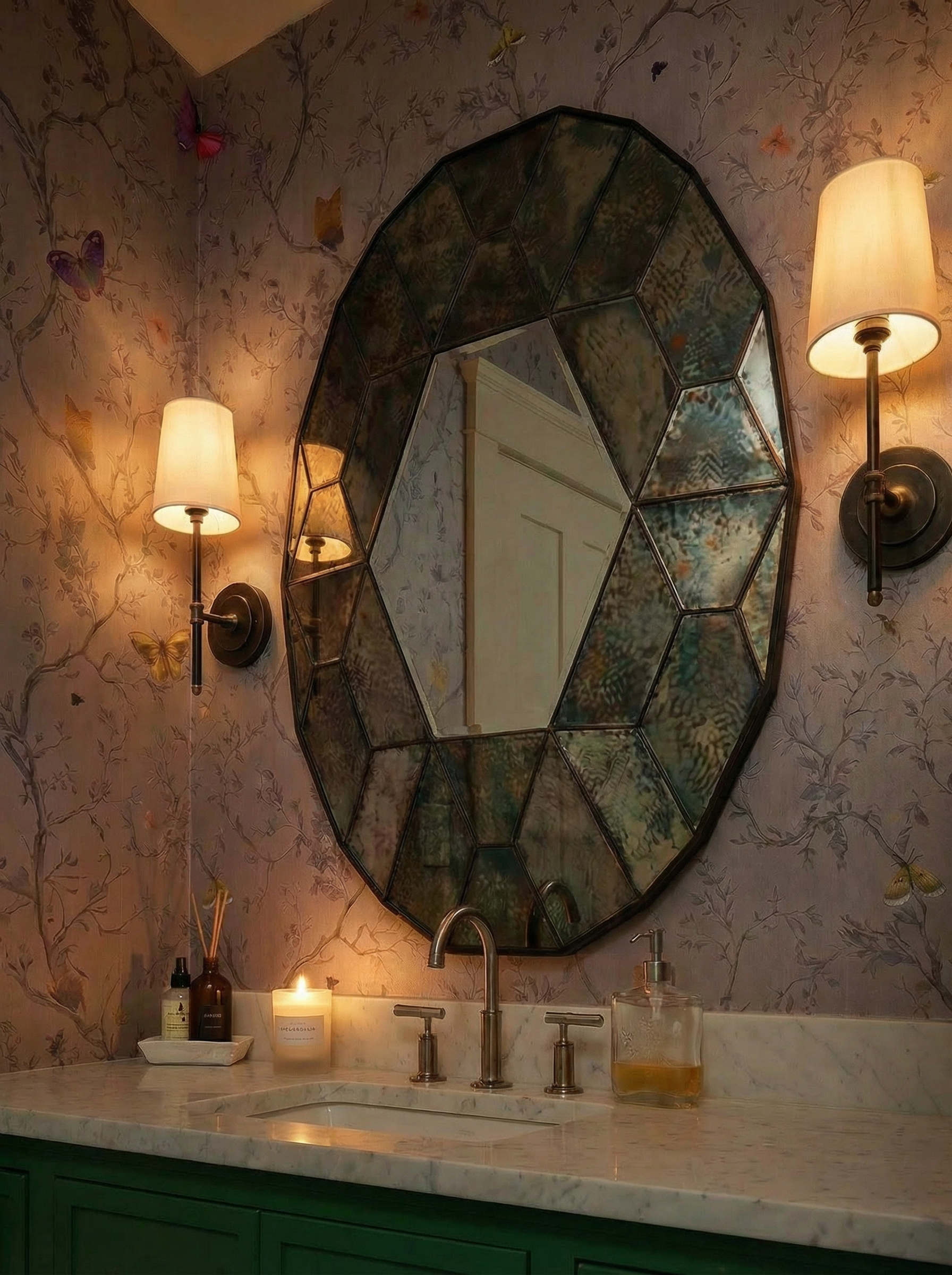 Decorative bathroom mirror with an irregular shape, mounted on wallpaper with butterfly and vine pattern, flanked by two wall sconces with lampshades, above a marble countertop with a gold soap dispenser, a candle, and small bottles.