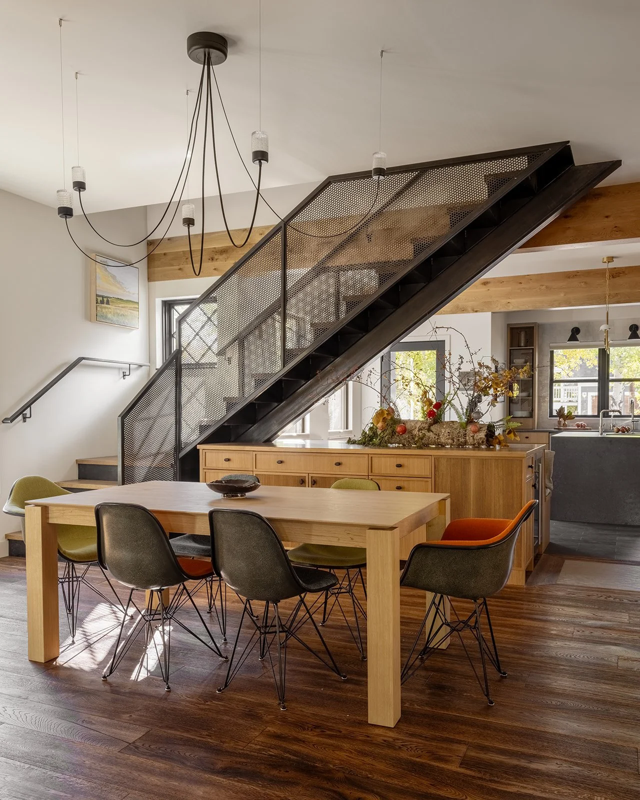 Modern dining room with wooden table, six mixed color chairs, wood flooring, and a staircase with black railing leading upstairs. Large windows, minimalistic decor, and a chandelier hanging from the ceiling. Bozeman, Montana, Remodel.
