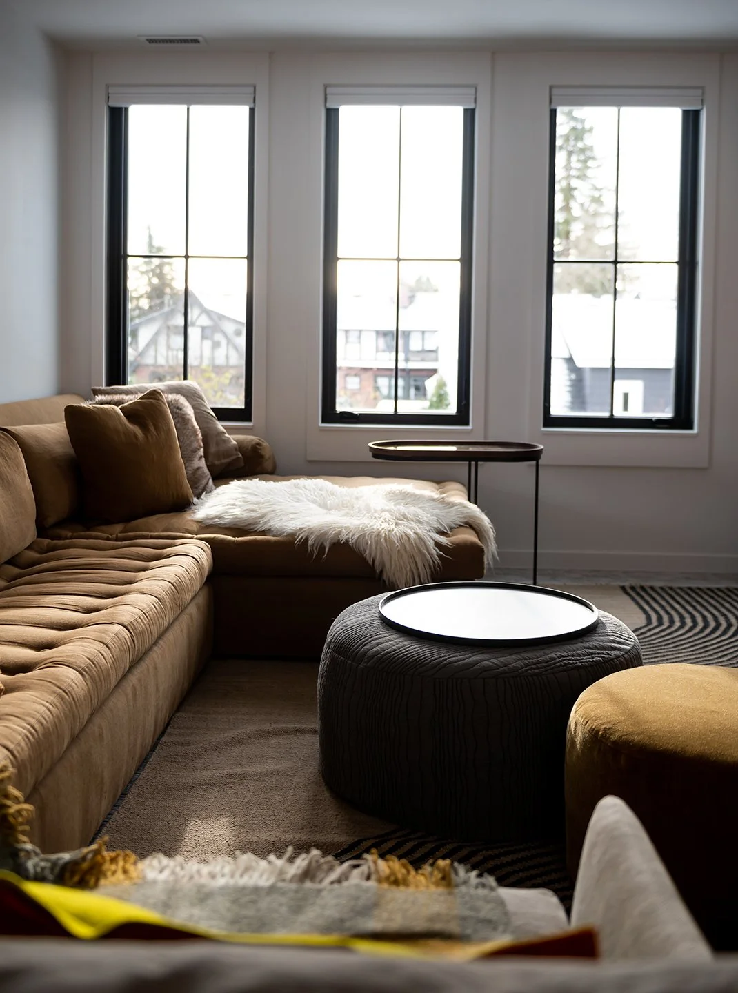 Living room with large windows, a beige sectional sofa with pillows, a white furry throw, a small round table, and a modern ottoman with a tray on top. Seattle, Capitol Hill, Remodel.