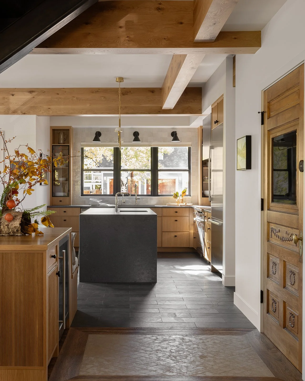 Modern kitchen with wooden cabinets, black window frames, a central dark stone island, wooden ceiling beams, and gray flooring. Features natural light from large windows.