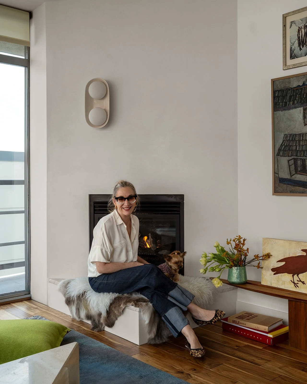A woman sitting on a white fur-covered bench in front of a fireplace with a dog, inside a modern living room with artwork and a vase of flowers. Seattle, Belltown, Condo, Remodel.