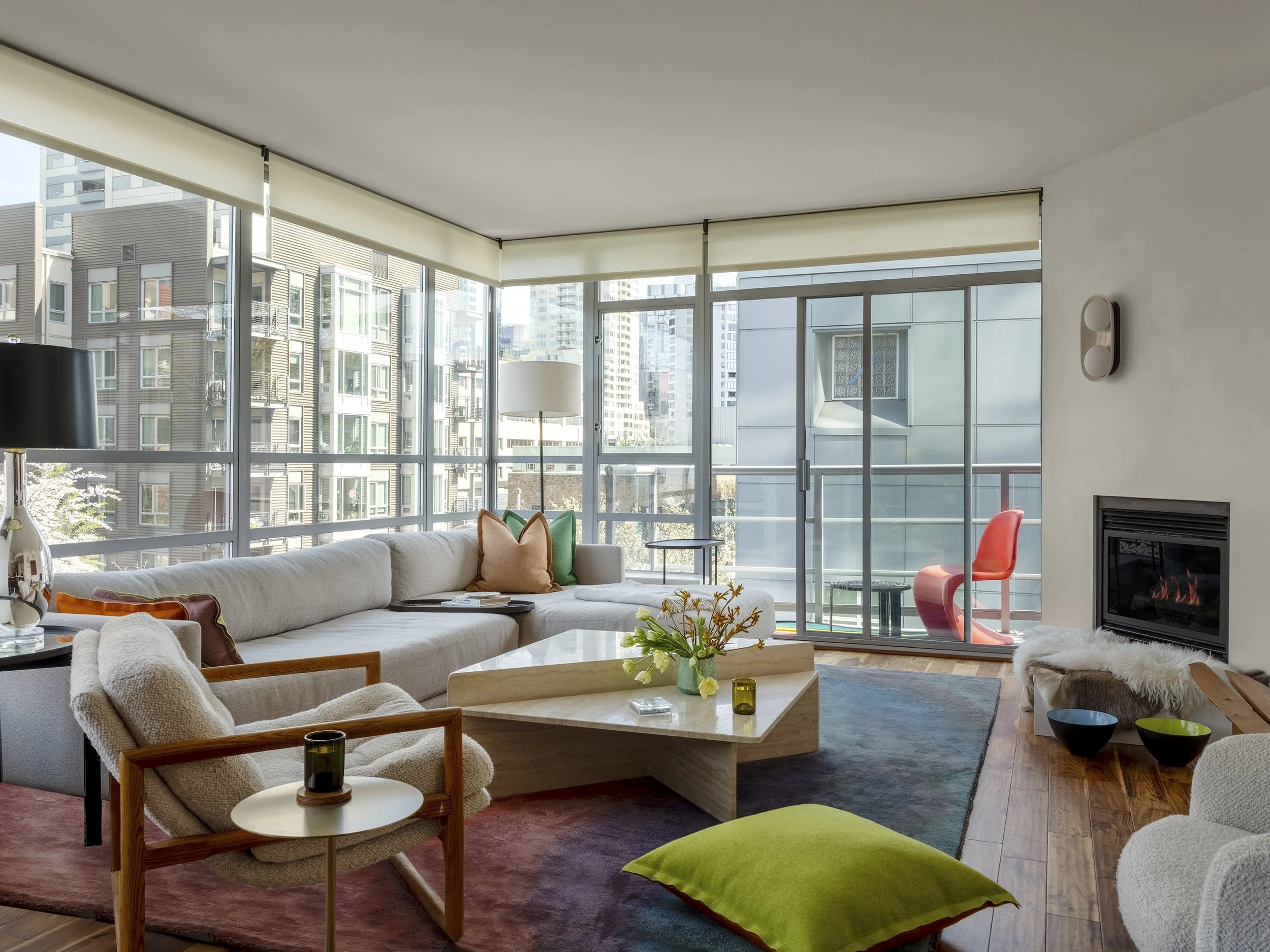 Bright living room with large windows, white sofa with colorful pillows, armchair with blanket, modern coffee table with flowers, and a fireplace. Seattle, Belltown, Condo, Remodel.