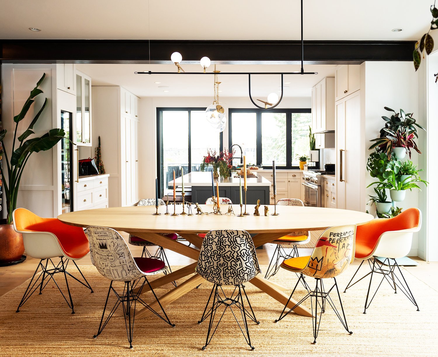 Modern dining room with an oval wooden table surrounded by colorful, eclectic chairs. The room features a bright and open kitchen with white cabinets and large windows. Indoor plants and contemporary pendant lights add to the decor.