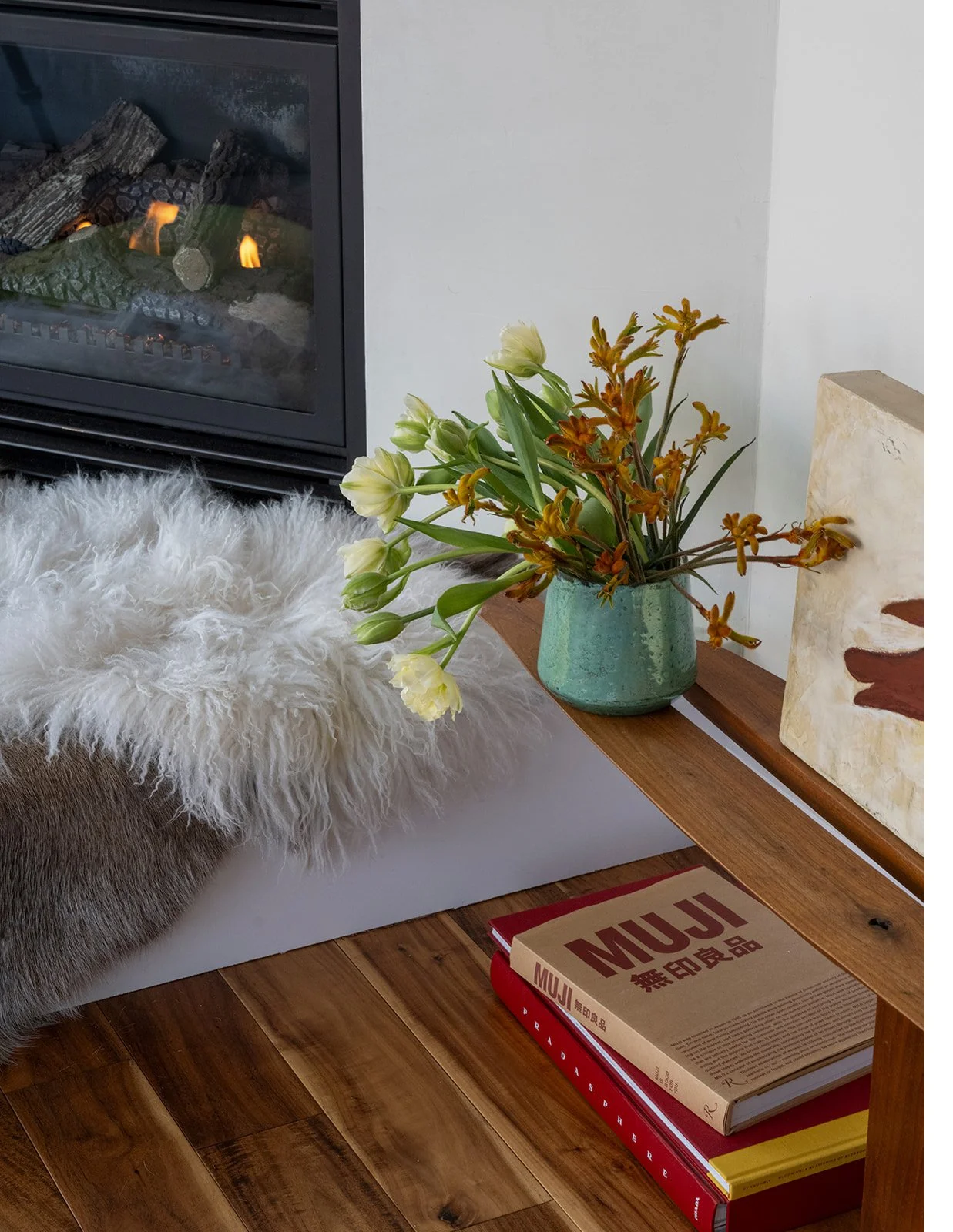 A vase with white and yellow flowers on a wooden shelf next to a painting, a pile of books, and a faux fur throw on the floor near a fireplace. Seattle, Belltown, Condo, Remodel.