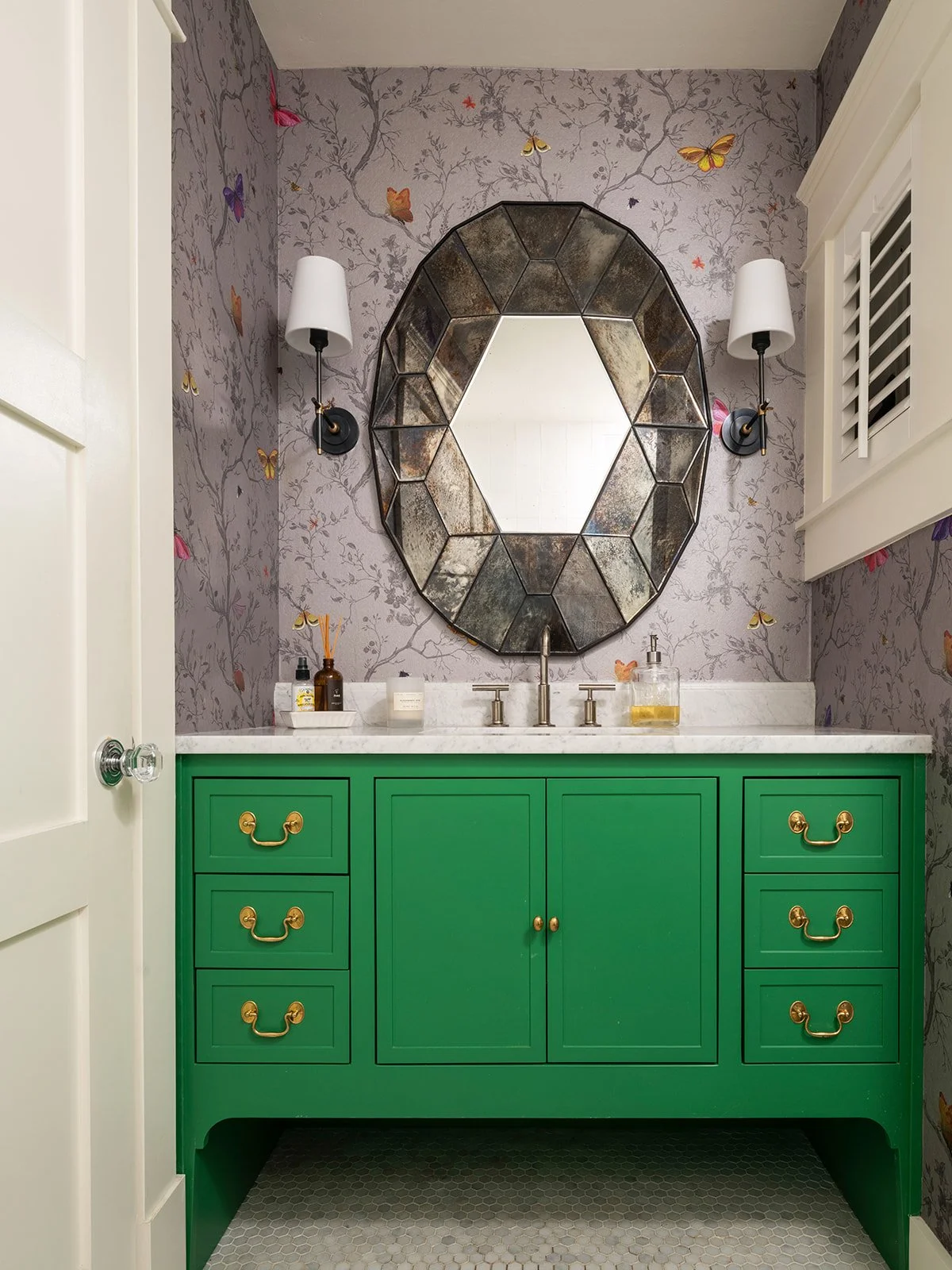 Green bathroom vanity with gold handles, a white marble countertop, and a decorative mirror above, flanked by two wall sconces, in a bathroom with butterfly-themed wallpaper. Seattle, Queen Anne, Basement, Remodel.