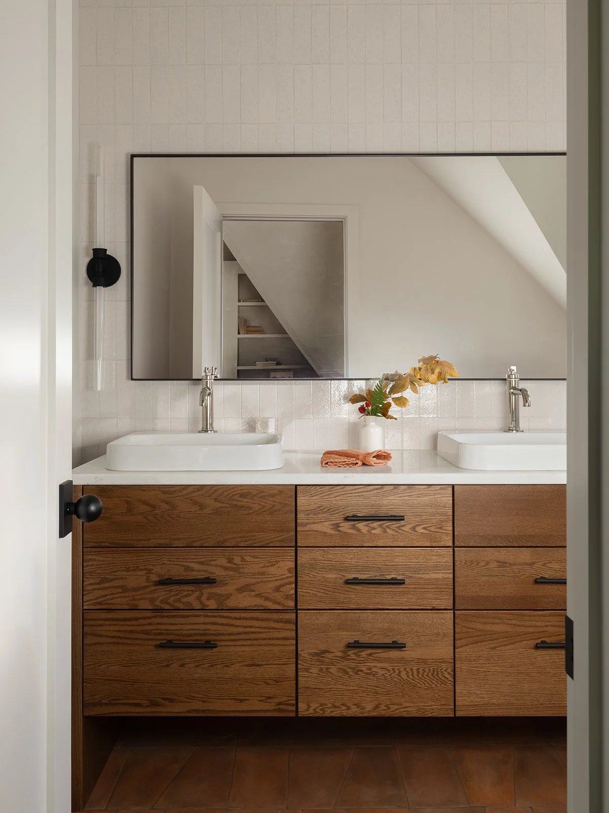 Bathroom vanity with a large mirror, two vessel sinks, a wooden cabinet, and a white countertop. Decorative foliage in a white vase and a folded towel are on the counter. Bozeman, Montana, Remodel.