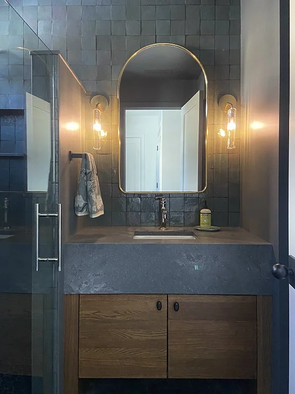 Modern bathroom with a dark tile wall and a wooden vanity. Features an oval mirror with gold trim, two wall sconces, and a towel hanging on a rail. A glass shower door is visible on the left.