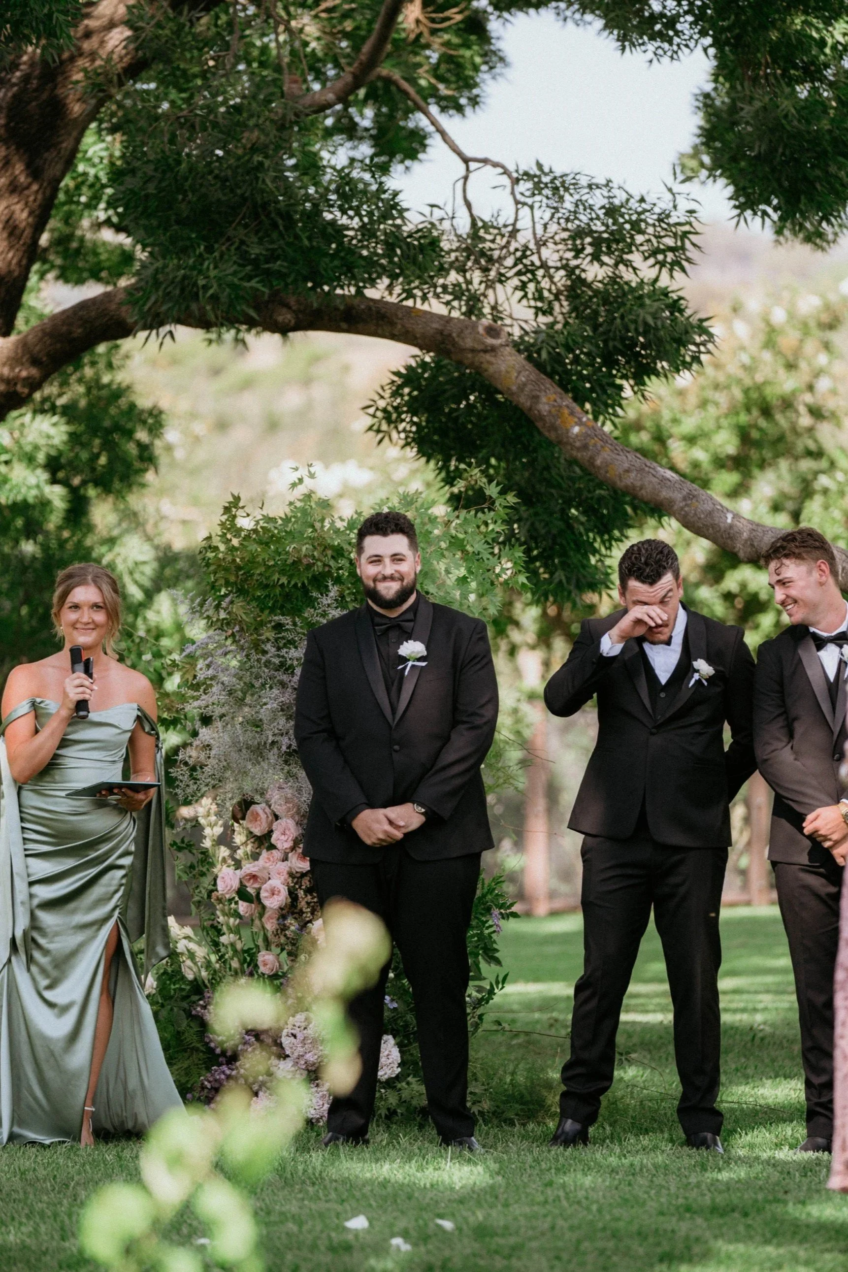 A wedding ceremony outdoors with four people standing in front of a floral arch and lush green trees. The man in the center is smiling, wearing a black tuxedo with a white boutonniere. The woman on the left is holding a microphone, wearing a green dress, and the other two men on the right are dressed in black tuxedos, one wiping his eye, possibly tearing up. The scene is decorated with pink and white flowers.