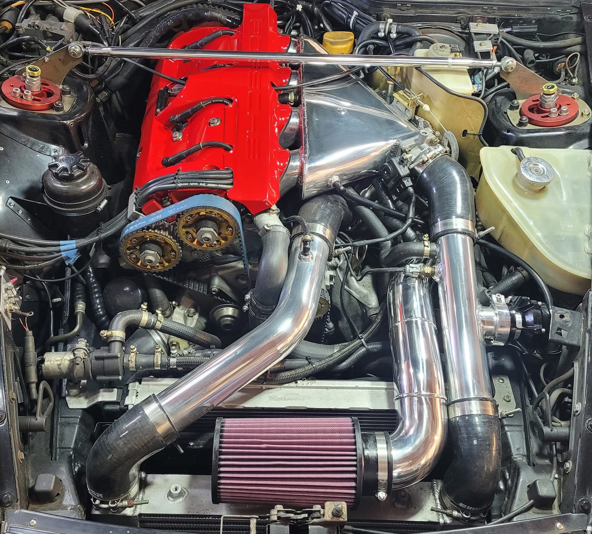 Modified car engine with a red valve cover, chrome intake pipes, and a large air filter.